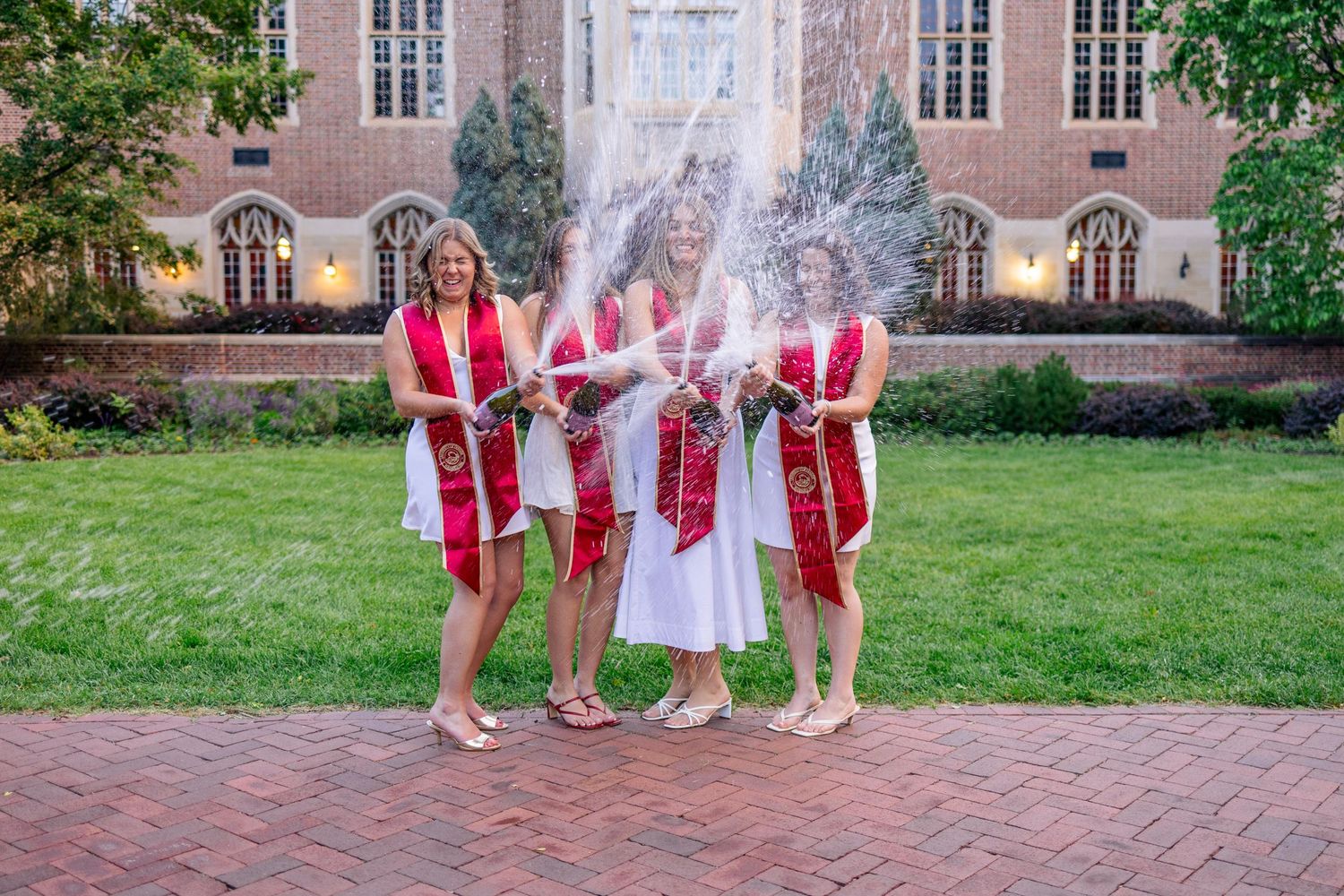 Three graduates in white dresses and red sashes celebrate with champagne spray outside a brick university building.