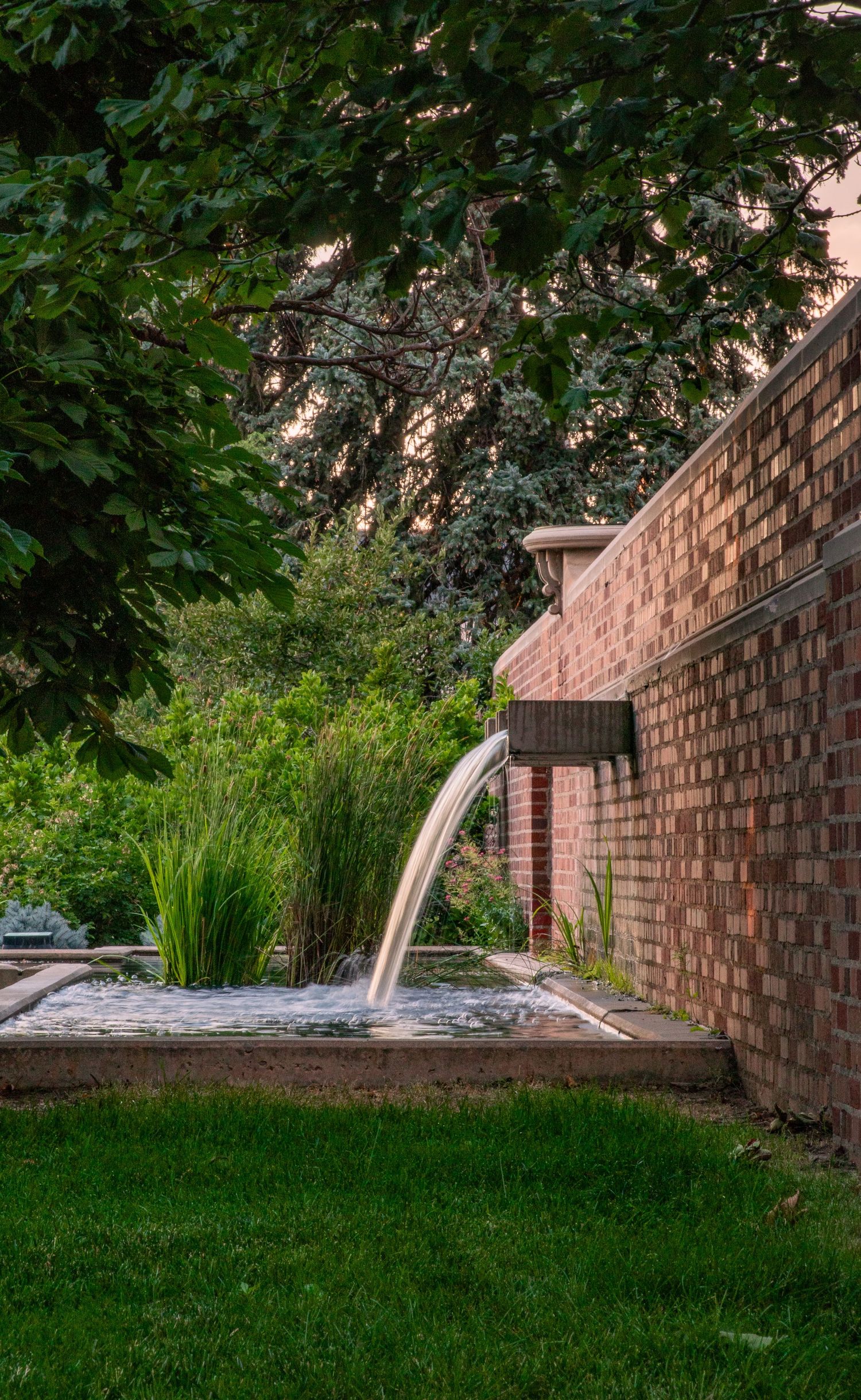Water drains from a series of copper spouts along a brick wall into a lush garden setting at dusk.