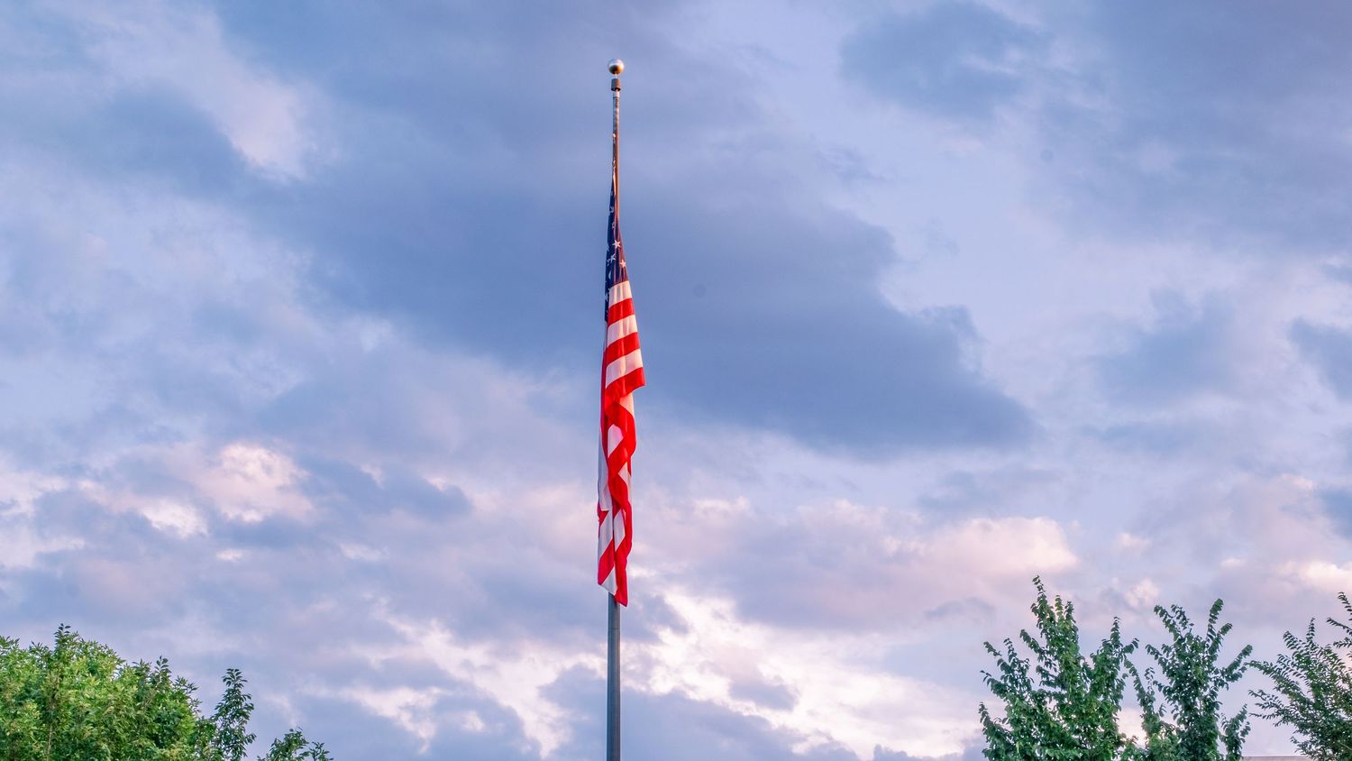 Six American flags wave proudly against a cloudy blue sky with trees visible in the background.