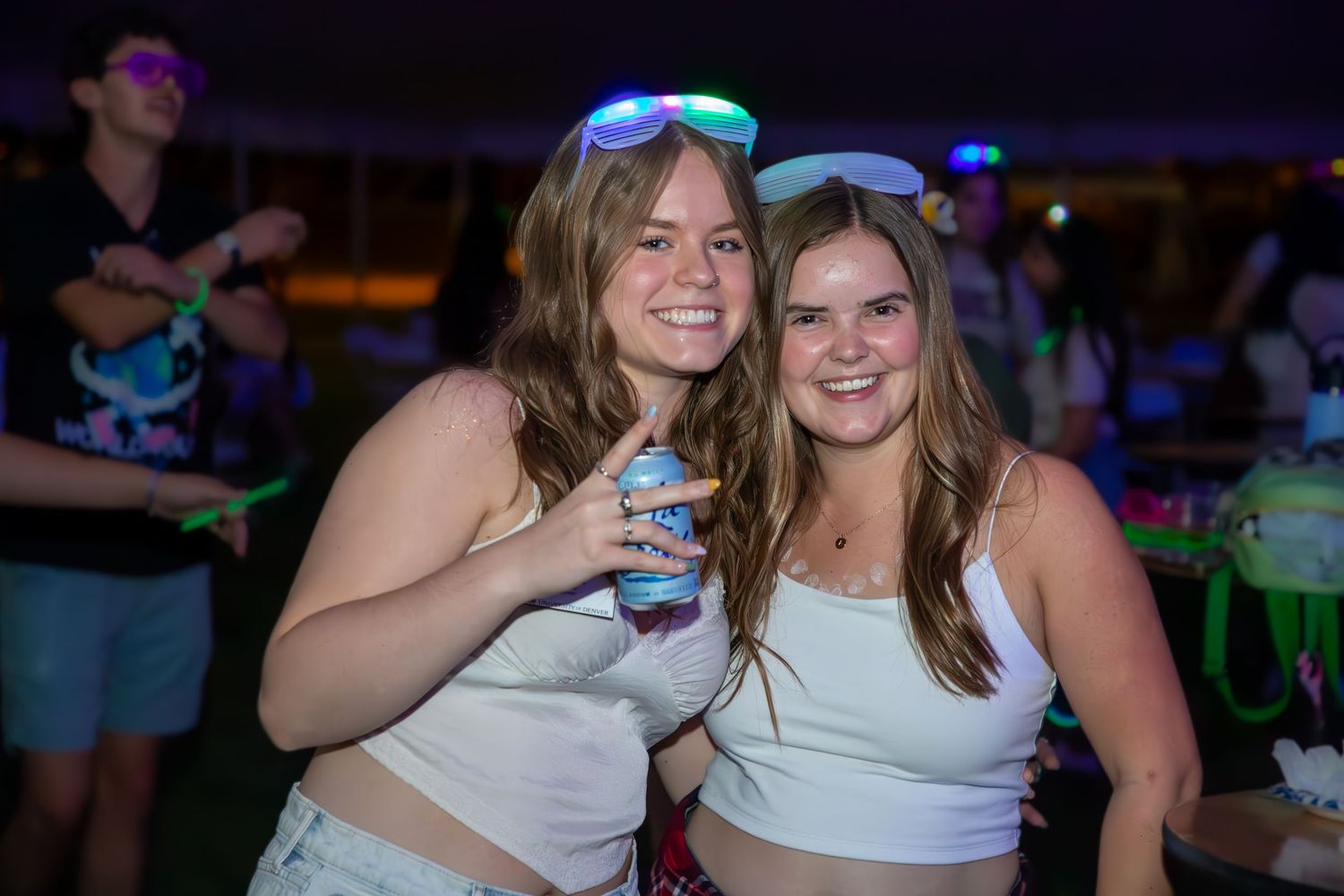 Two smiling young women posing together at a nighttime outdoor festival, one holding a drink.