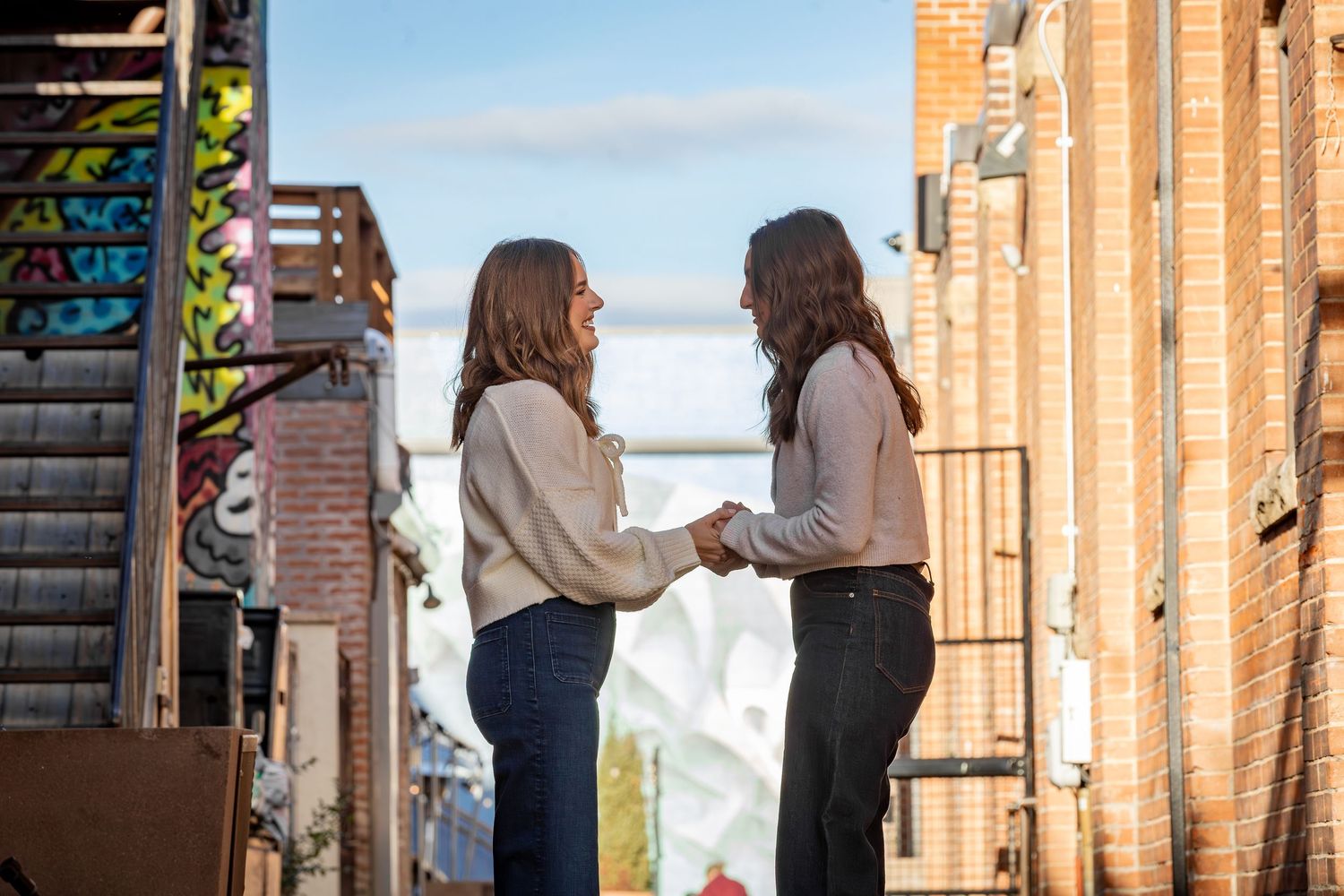 People walking through an urban alleyway with brick walls and sunlight casting long shadows on the ground.