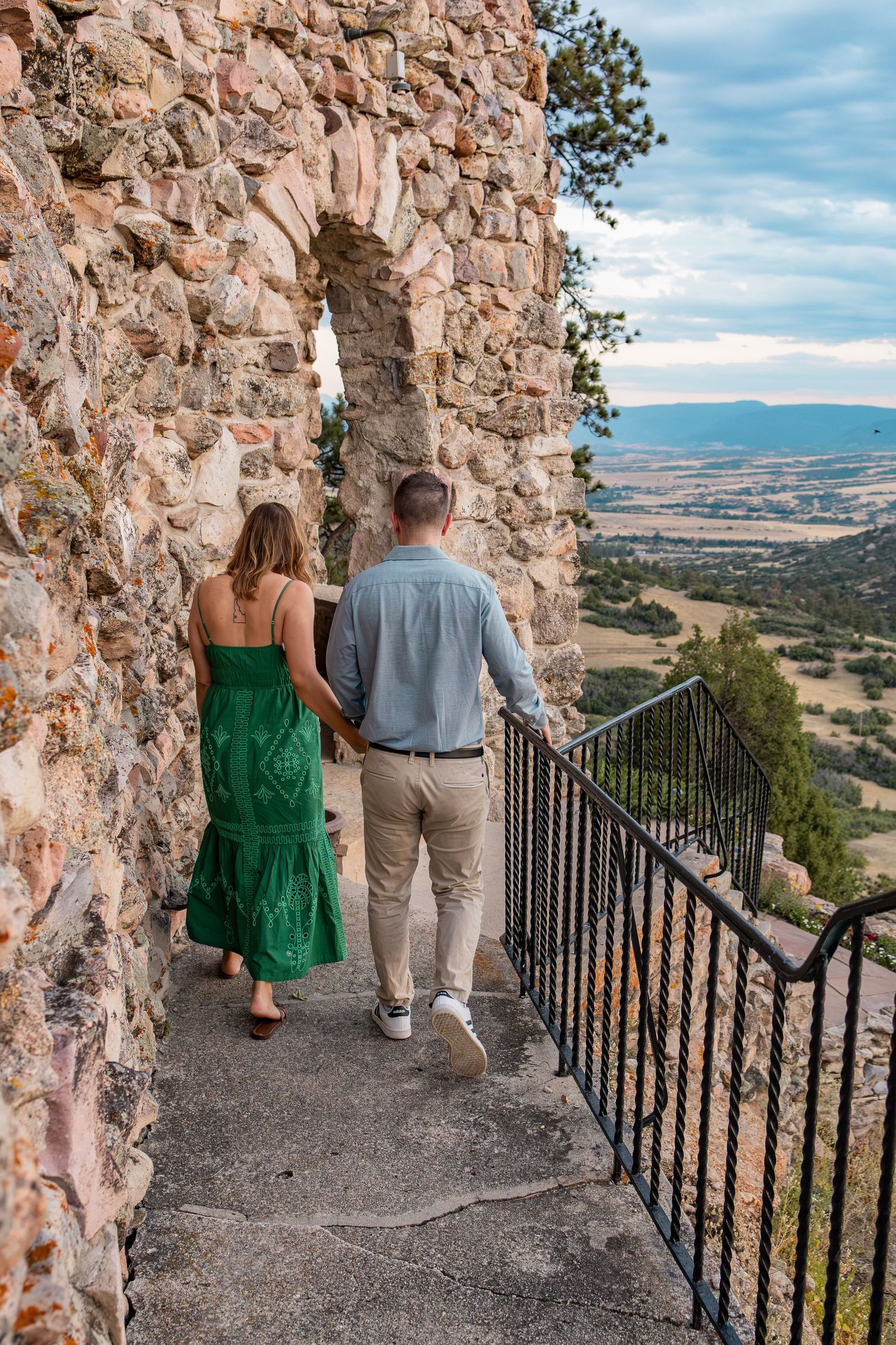 Couple holding hands walking along a stone castle pathway with scenic valley views in the background.