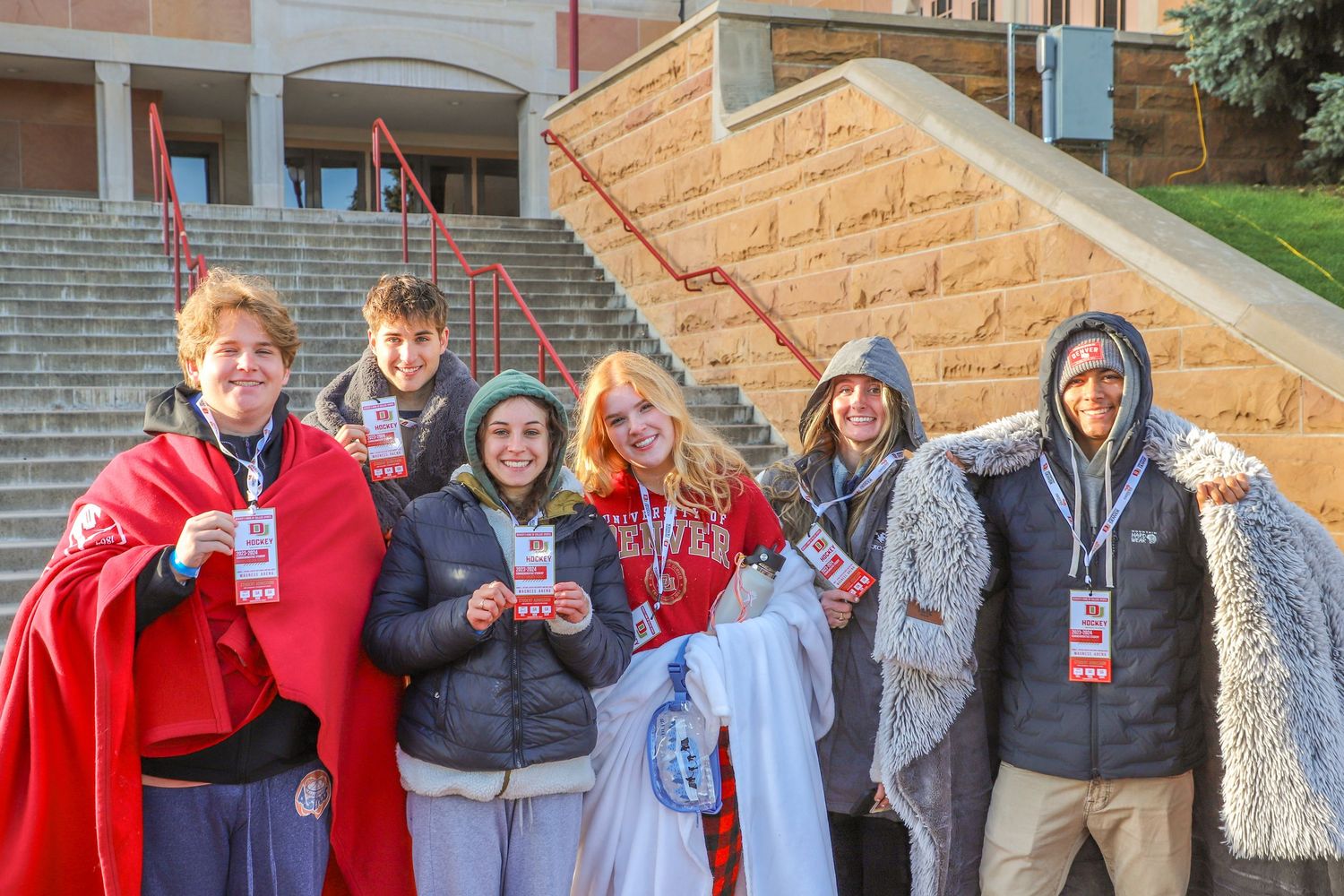 Group of six college fans bundled in blankets and jackets posing together outside a stadium on a cold game day.
