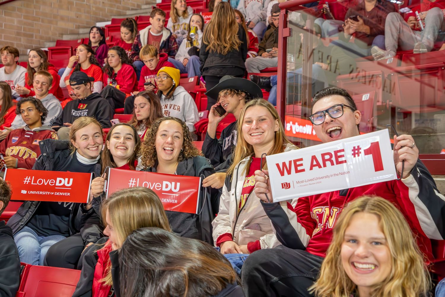 University of Denver students cheer at a hockey game holding #LoveDU and We Are #1 signs in red arena stands.