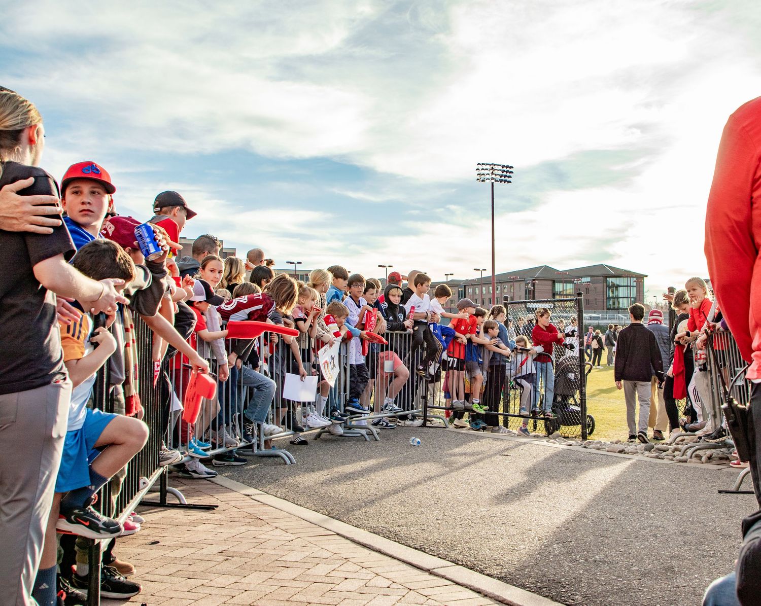 Large crowd of fans lines barriers outdoors at a red-themed sporting event.