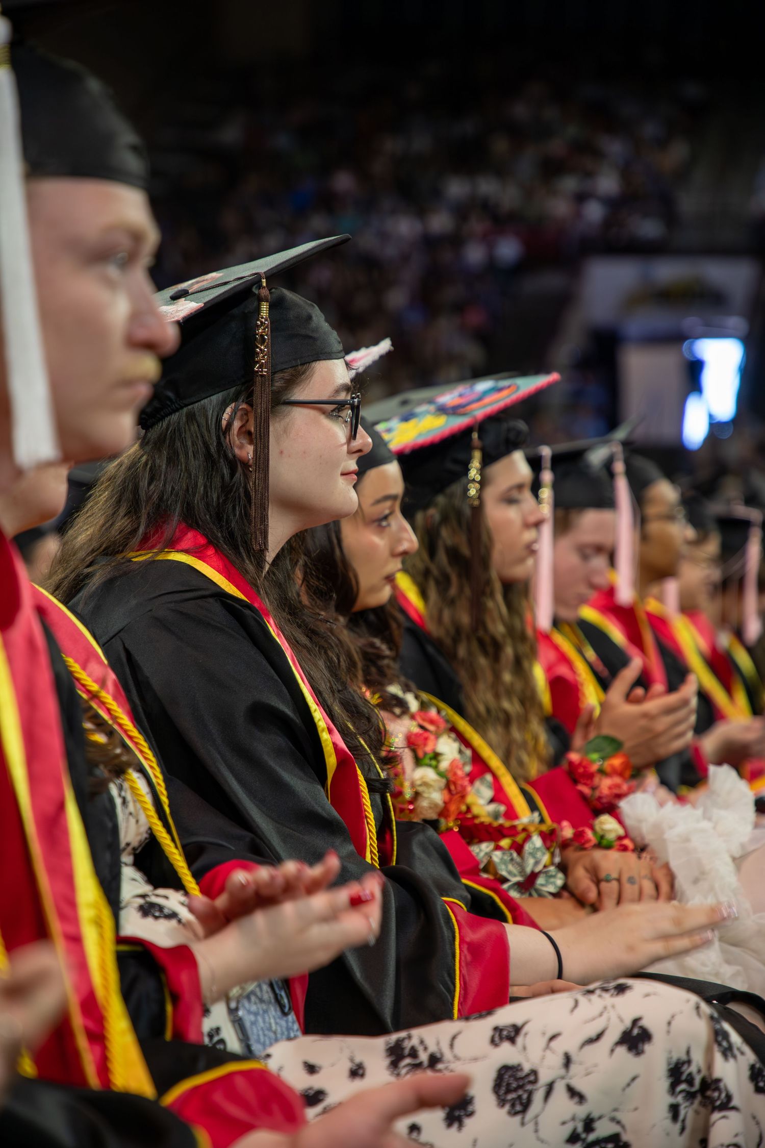 Graduates in black robes and decorated caps sit together during a college commencement ceremony.
