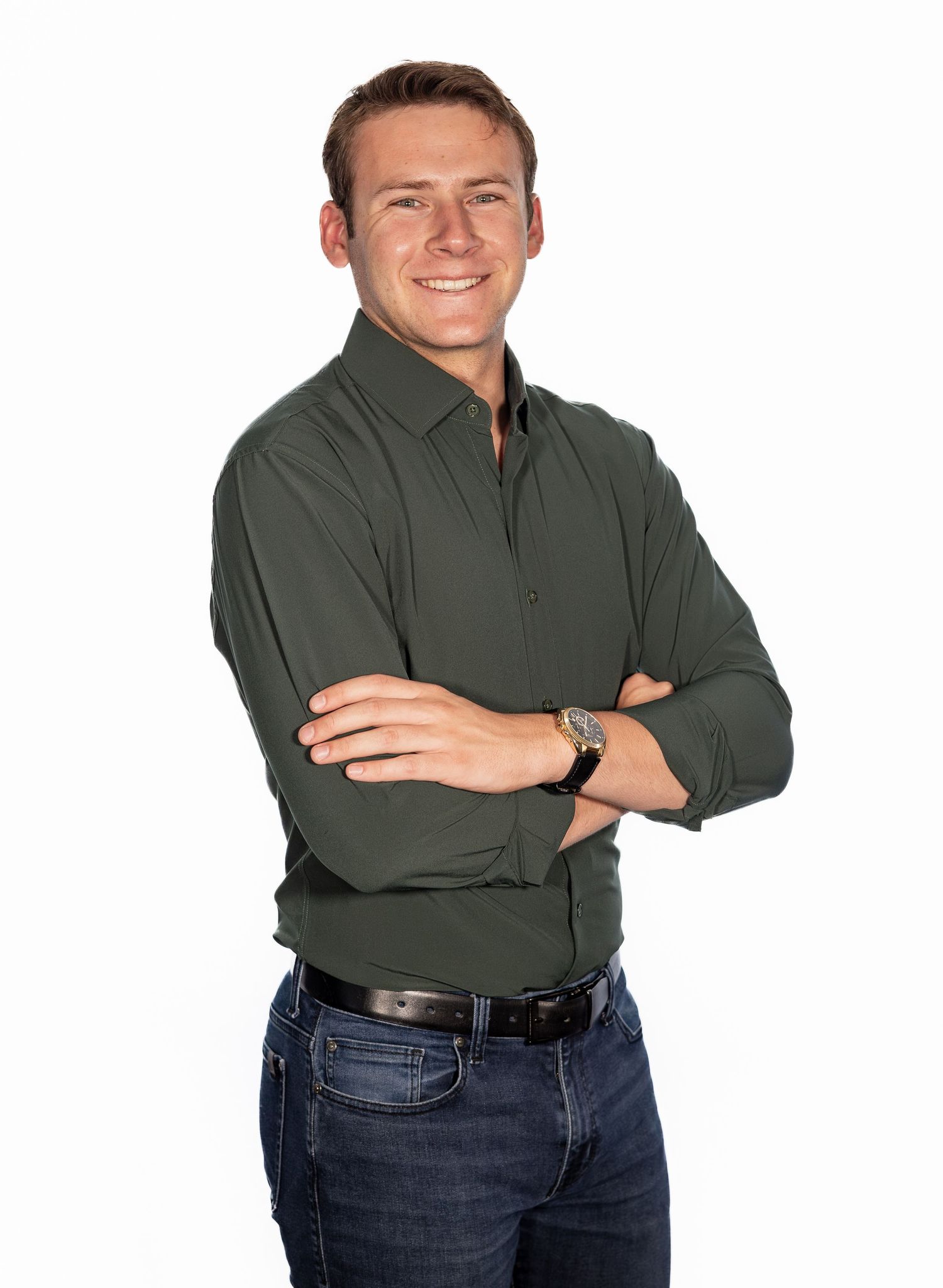 Smiling young man in dark green shirt with arms crossed, wearing jeans and a watch, posing against white background.