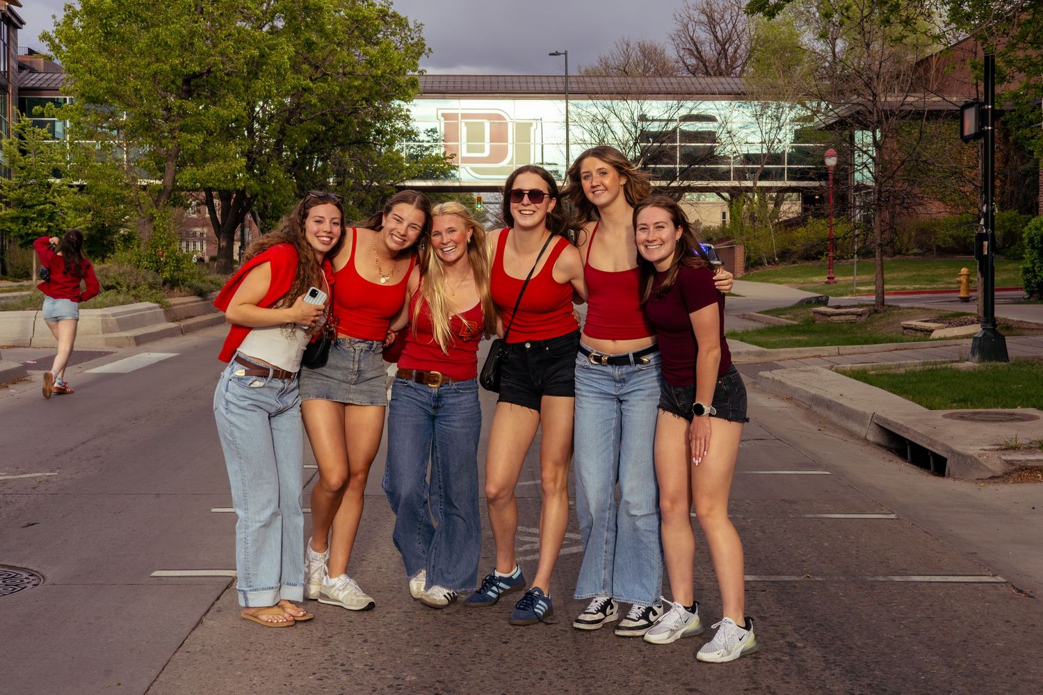 Six young women in red tops pose together outdoors on a college campus on a sunny day.