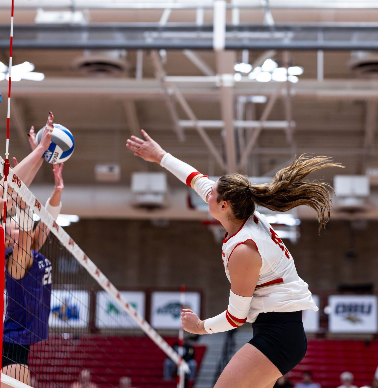 Female volleyball player spikes the ball over the net during an indoor match in a red and white uniform.
