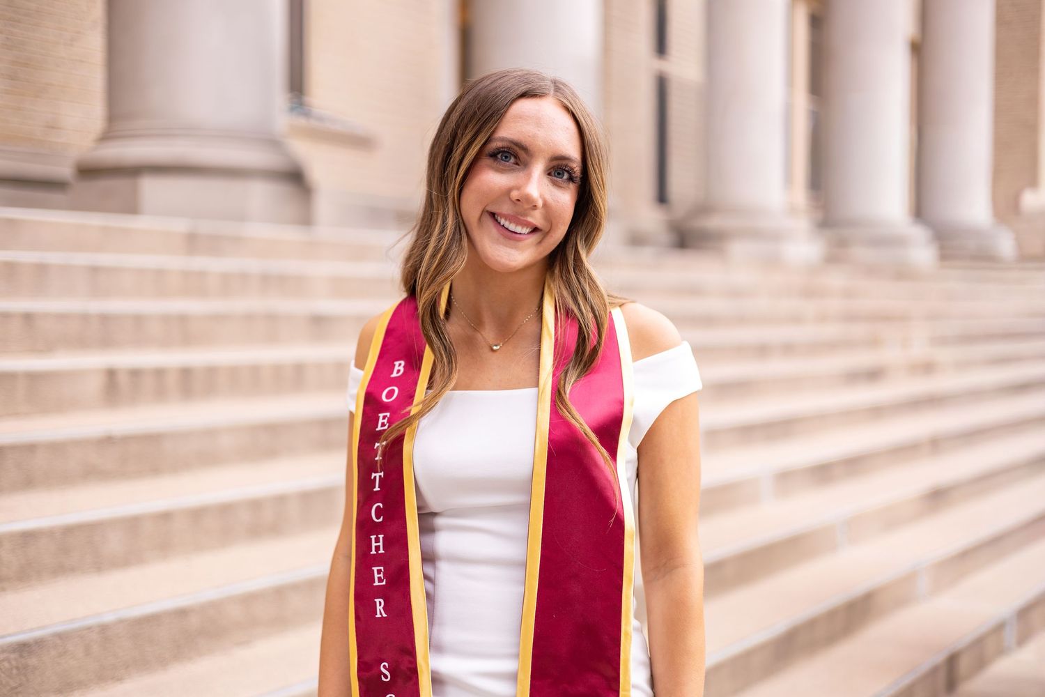 Smiling graduate wearing red and gold sash poses on stone steps with columns in background.