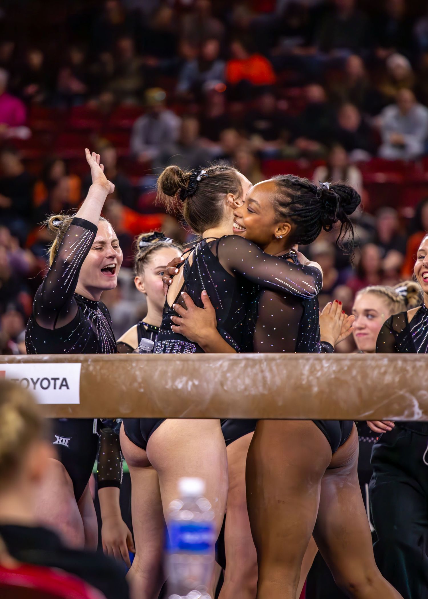 College gymnasts in black leotards celebrate together at a Big 12 gymnastics competition in a packed arena.