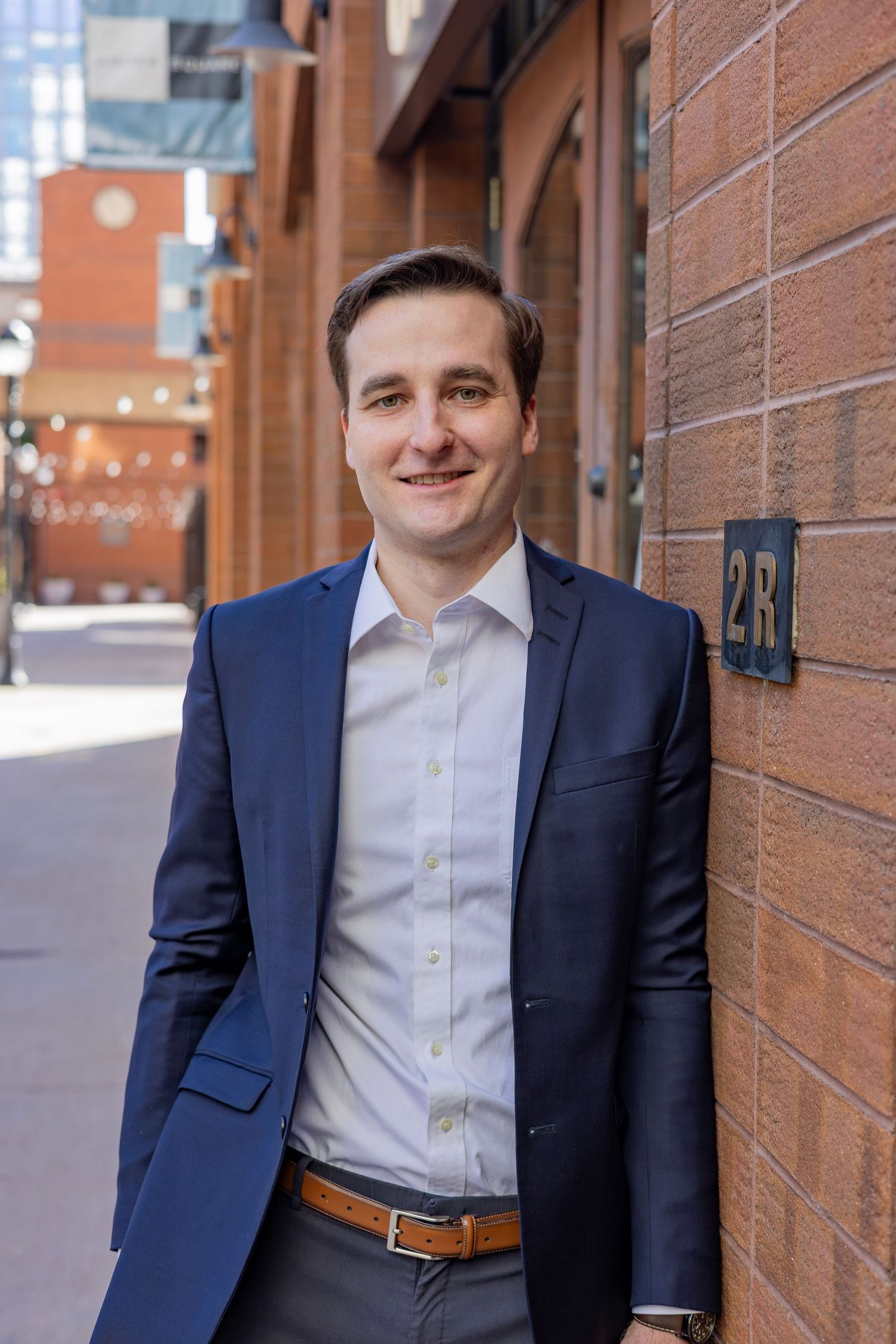 A smiling young professional man in a navy blue suit leans against a brick wall on an urban street.