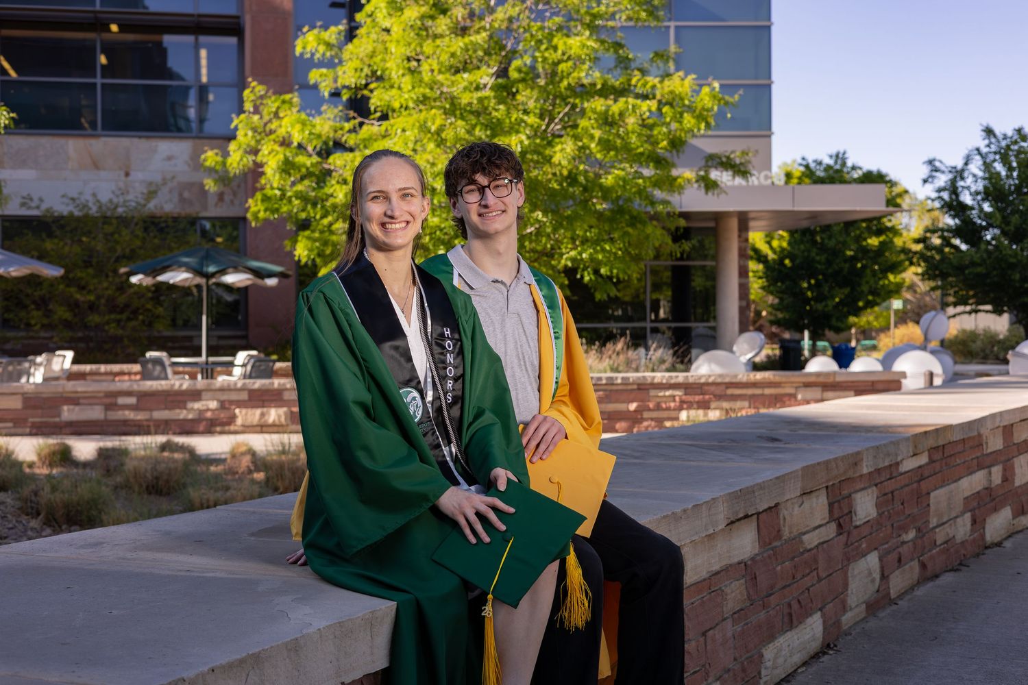 Two graduates in green and gold robes smile together outdoors at a university campus ceremony.