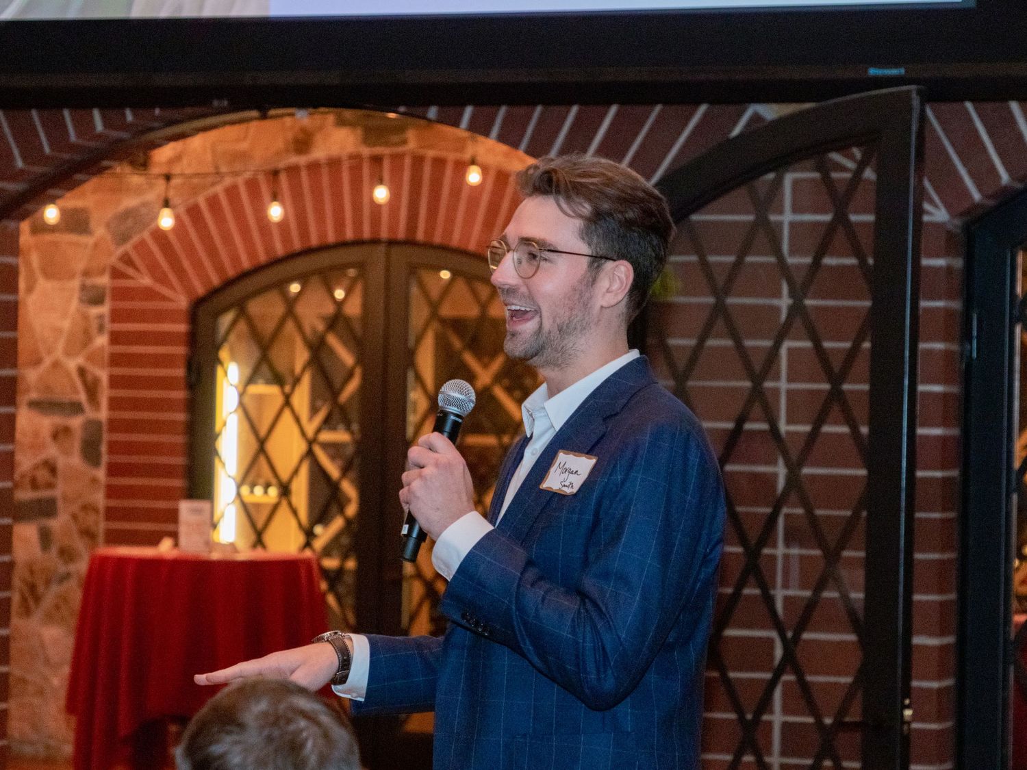 A speaker in a navy suit holds a microphone while presenting at an event in a brick-walled venue with warm lighting.