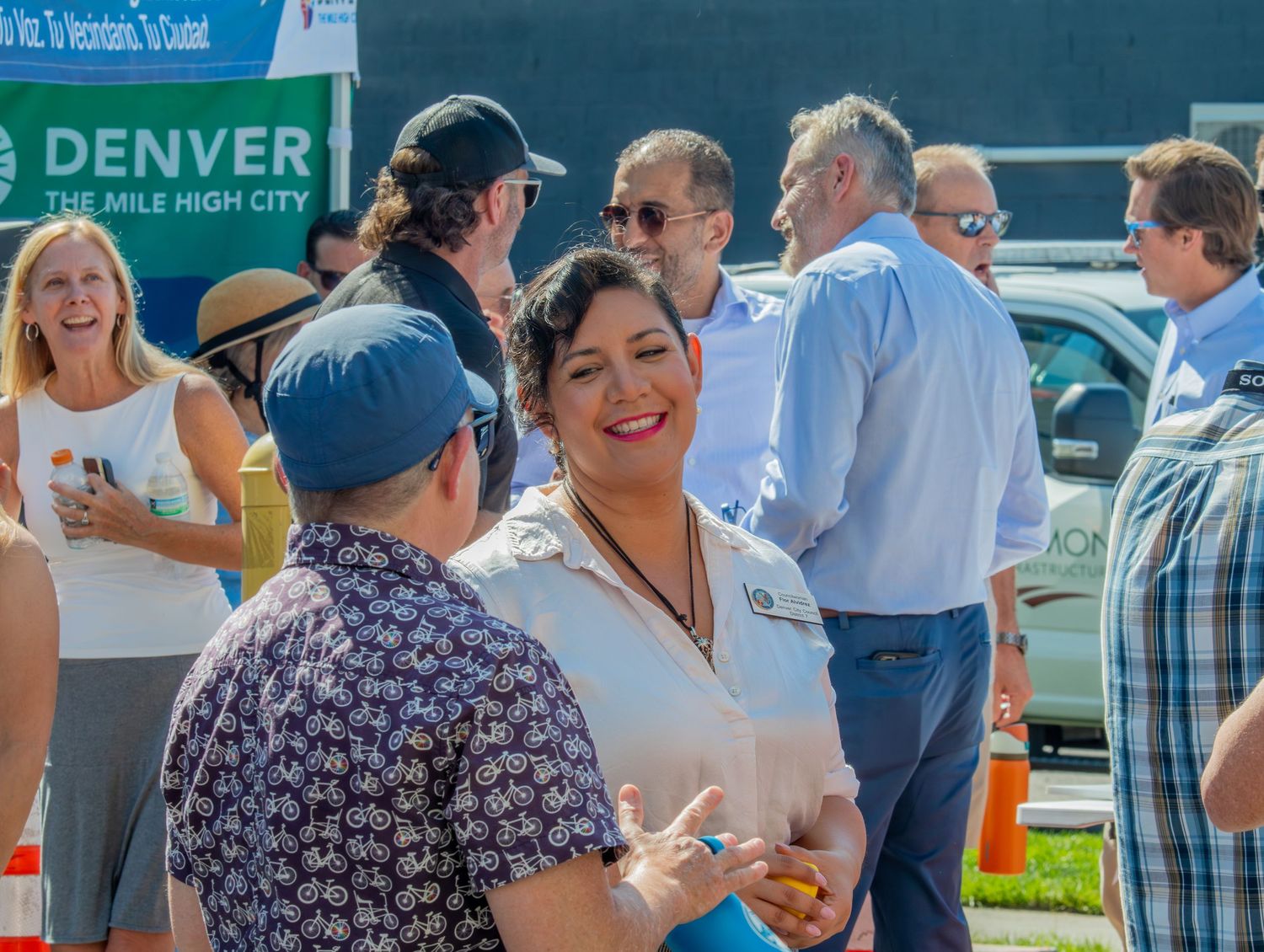 People gather at an outdoor event in Denver with food trucks and vendor booths visible in the background.