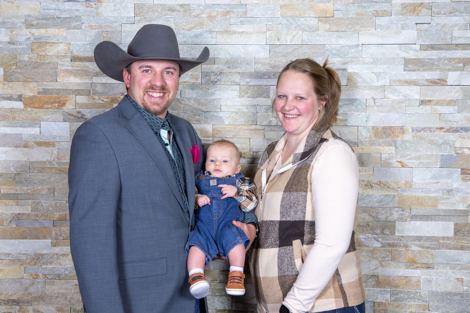 A family portrait against a stone wall backdrop featuring western attire and a small baby in overalls.