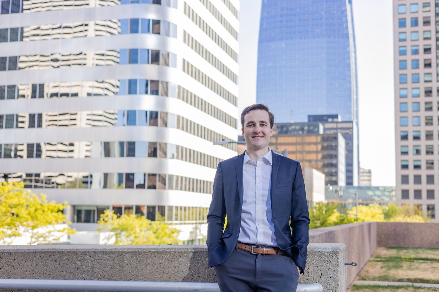 Young professional man in navy suit smiling outdoors with modern city office buildings in background on sunny day.