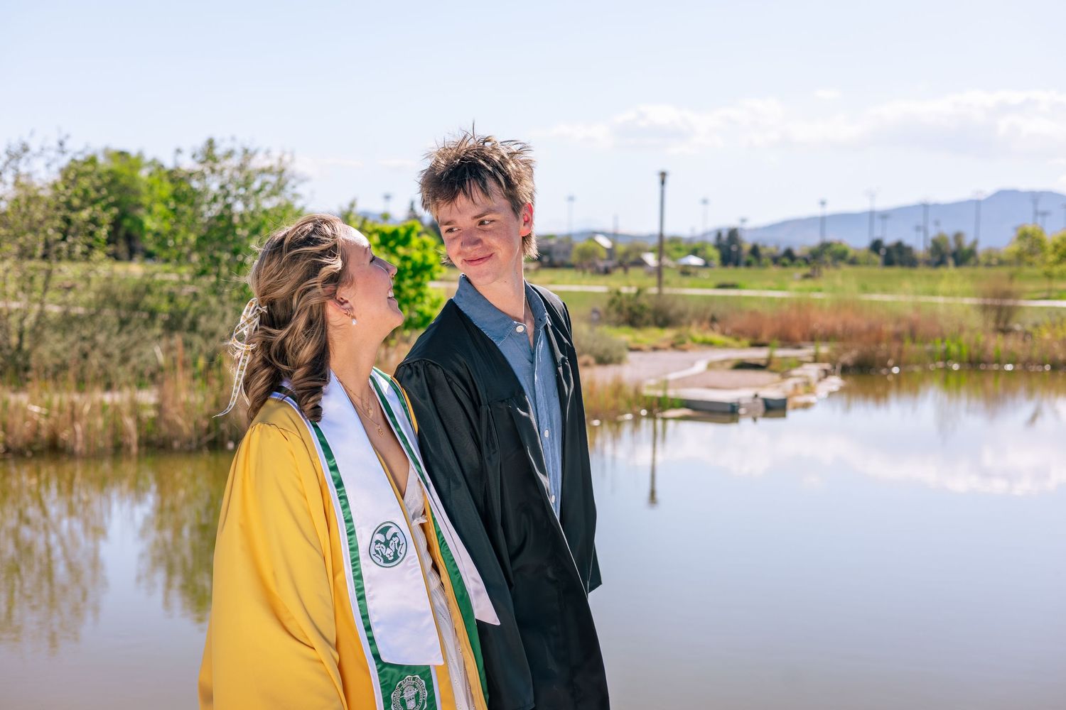 Two graduates in caps and gowns smile at each other near a reflective pond with mountains in the background.