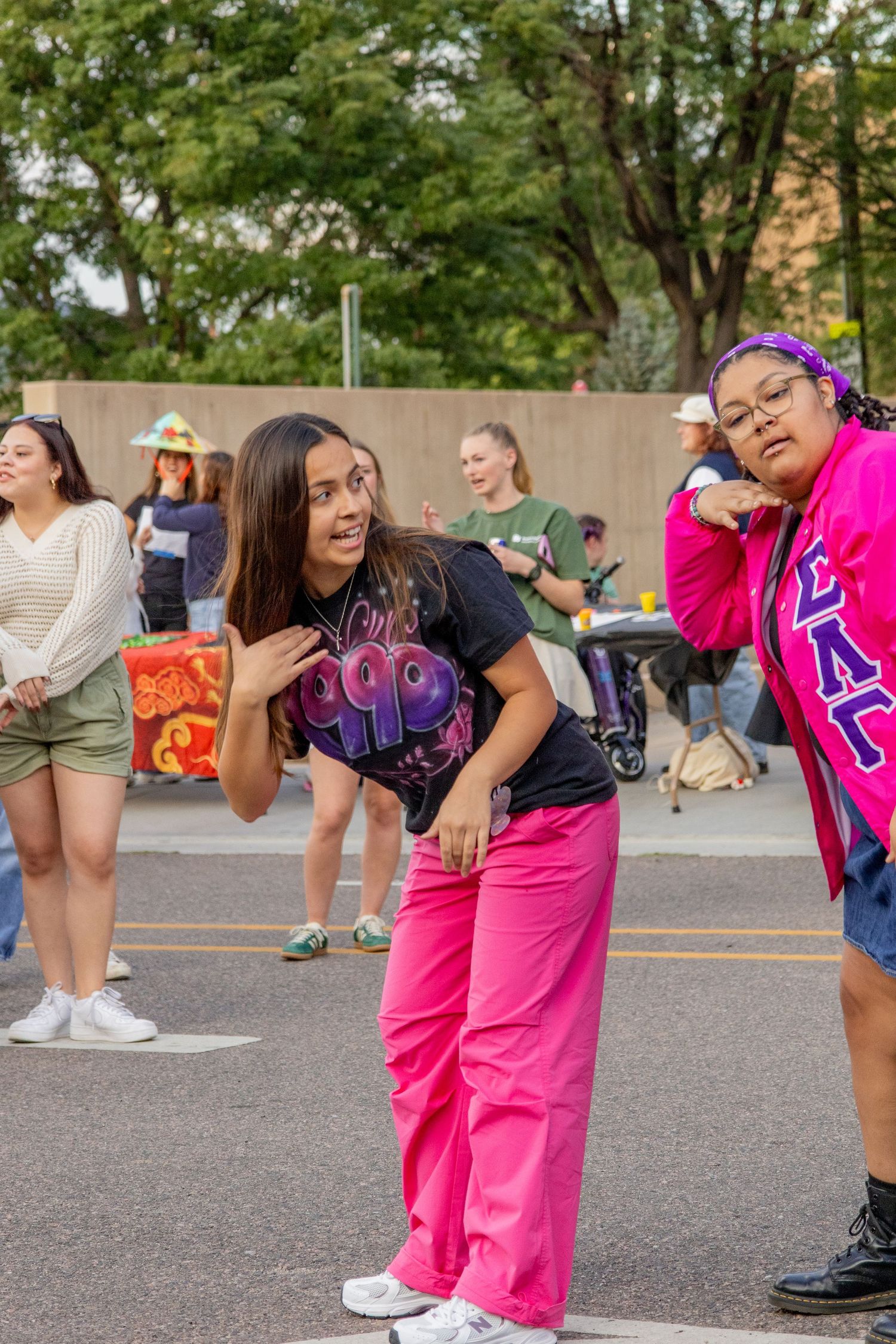 Two young women in pink pants and Greek letters shirt dance outdoors at a campus event with crowd behind them.