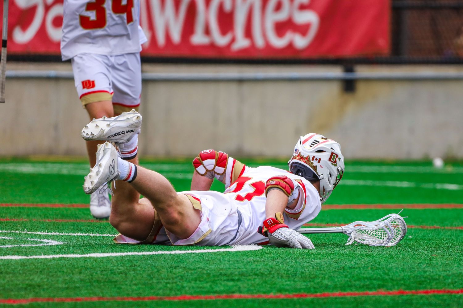 College lacrosse player in white uniform slides across green turf field during game action.