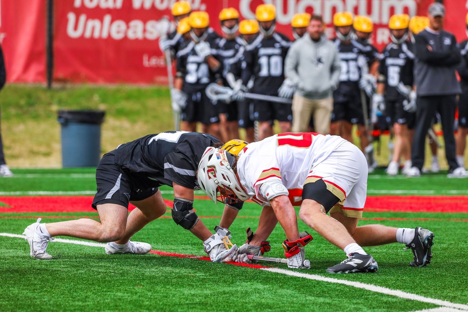 Two lacrosse players face off at midfield during a competitive college lacrosse game on a turf field.