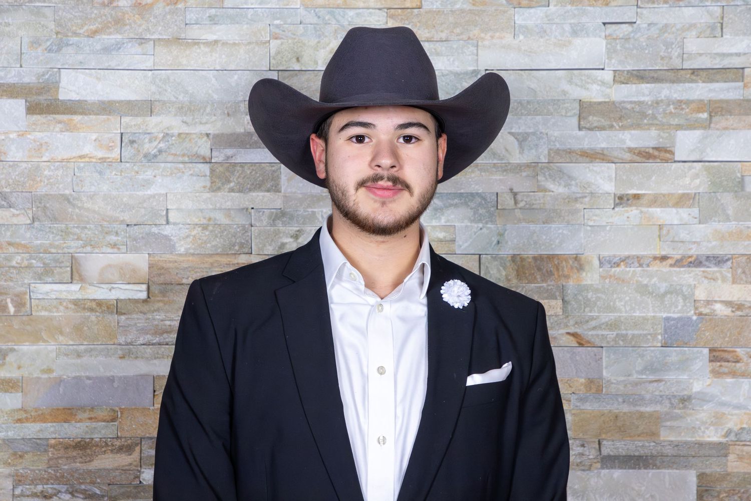 A formal portrait of a person in a black cowboy hat and suit against a textured stone wall.