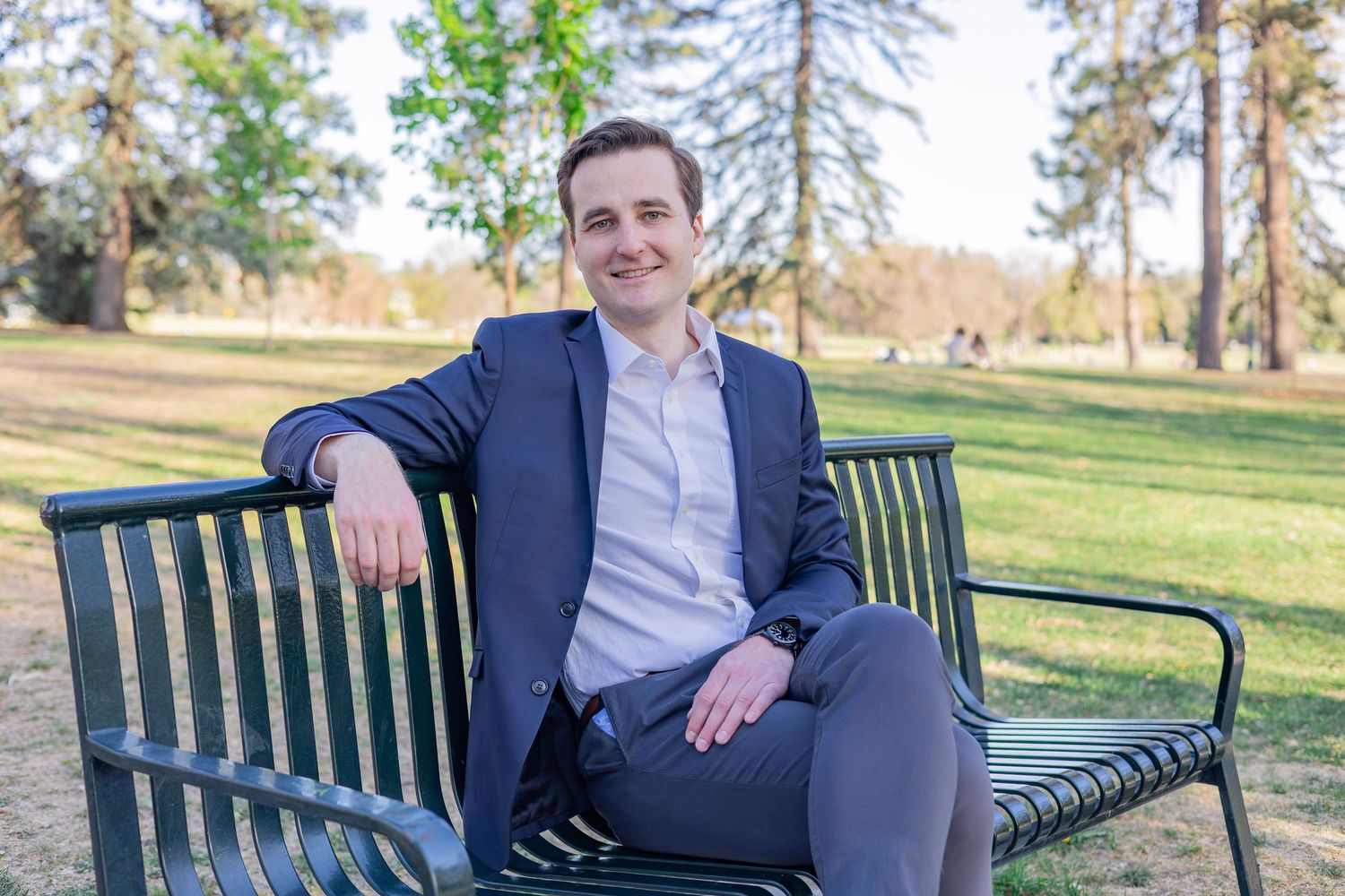 Young professional man in navy blazer sitting on park bench surrounded by lush green trees on a sunny day.