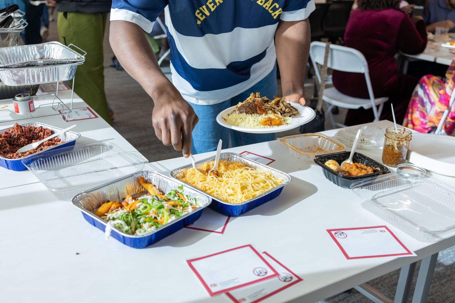 Person serving food at a buffet table with trays of mac and cheese, salad, and other dishes at an outdoor event.