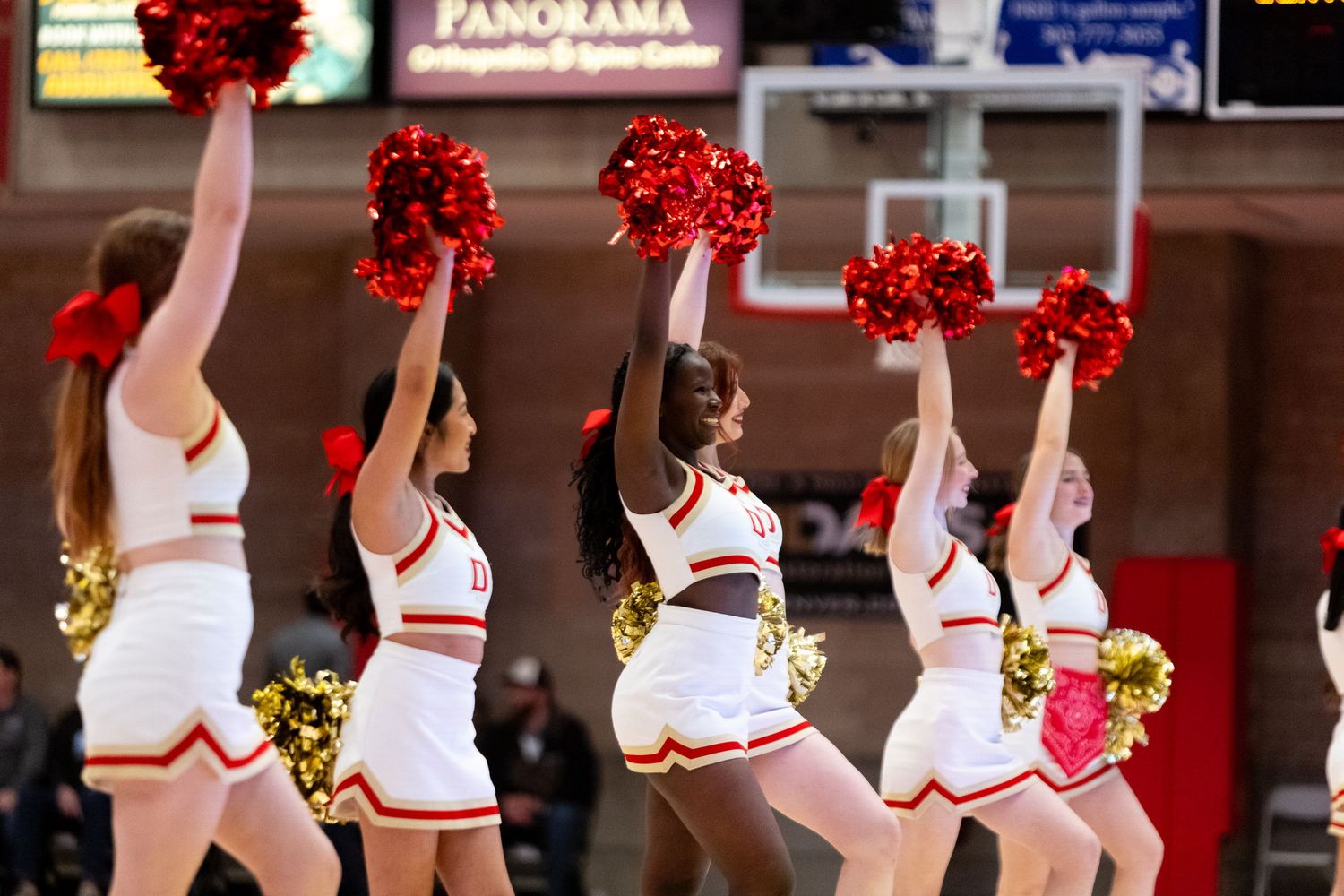 Cheerleaders in red and white uniforms perform with red and gold pom-poms at an indoor basketball game.