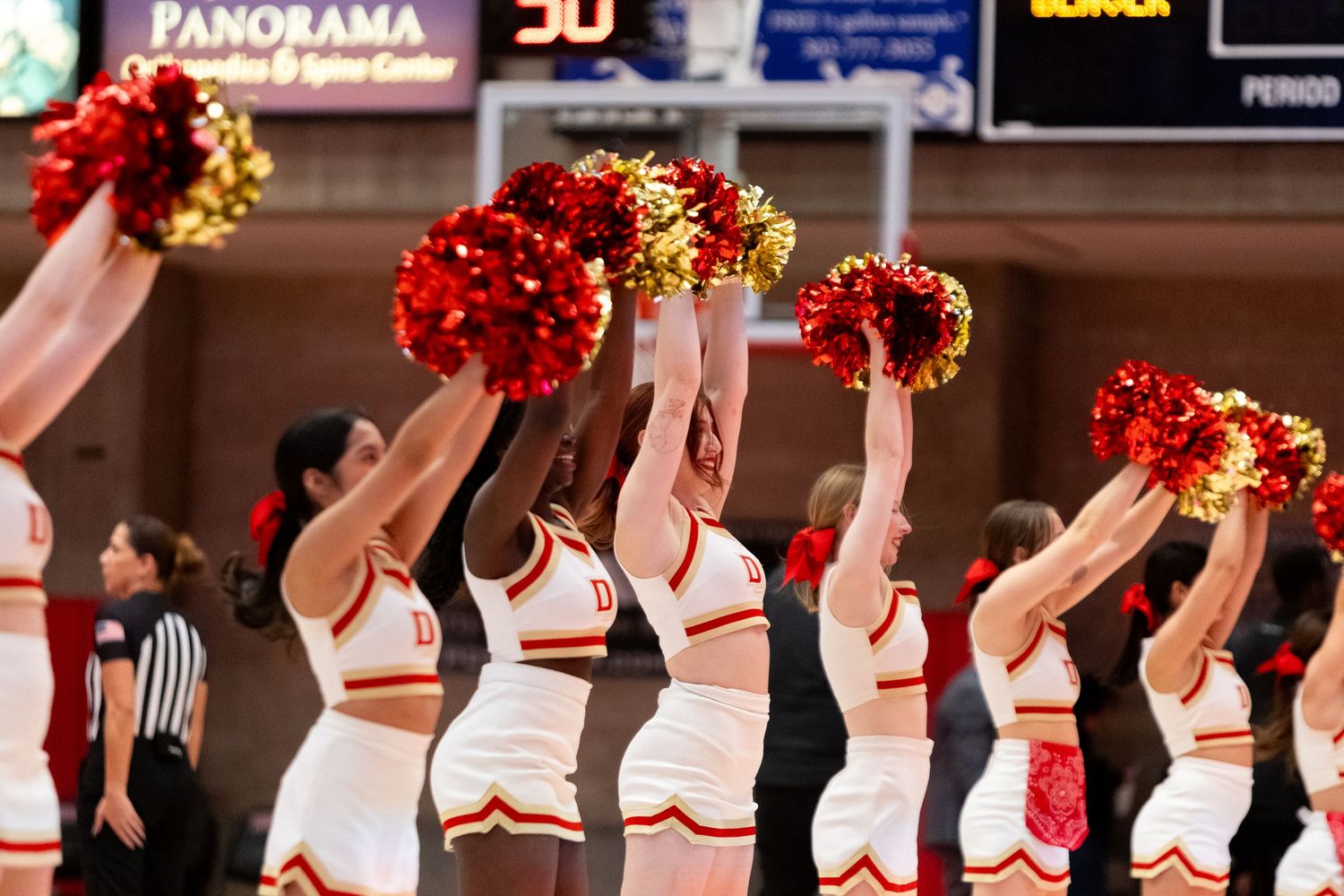 Cheerleaders in red and white uniforms perform synchronized dance routine with pom poms during basketball game.