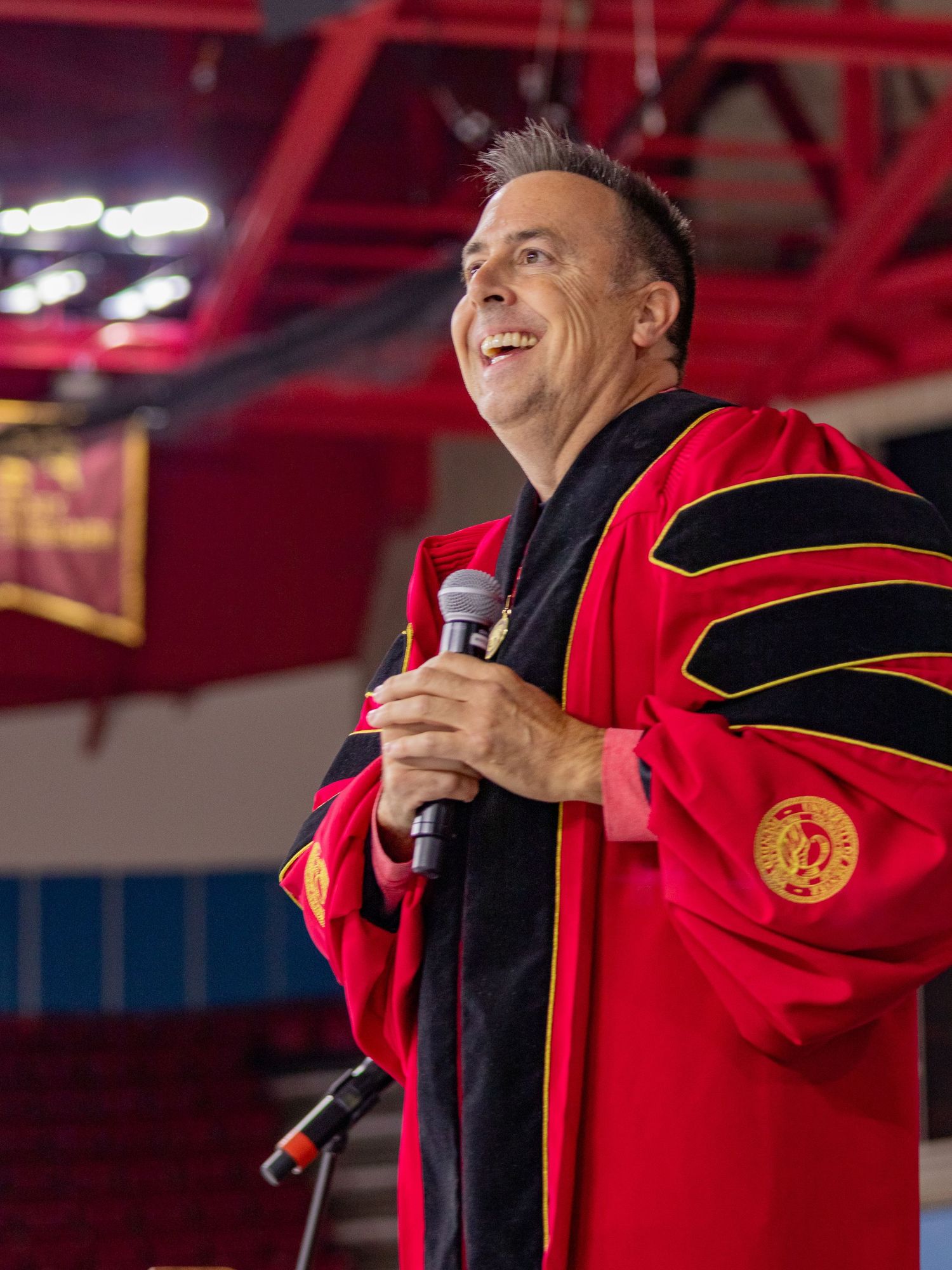 Man in red doctoral graduation regalia speaks at commencement ceremony holding microphone on stage.