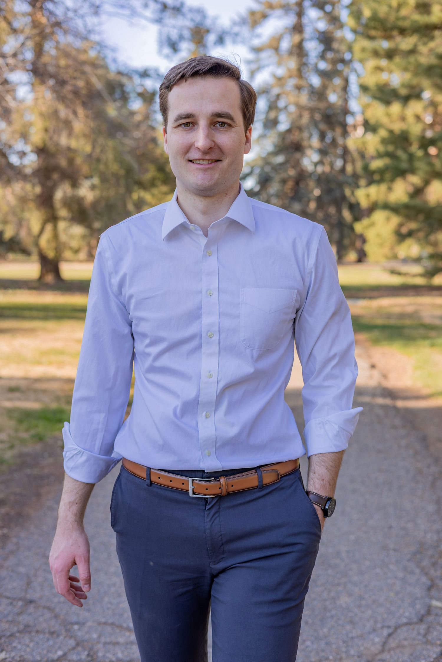 Man in white dress shirt and gray pants walks confidently along a tree-lined path in a sunny park.