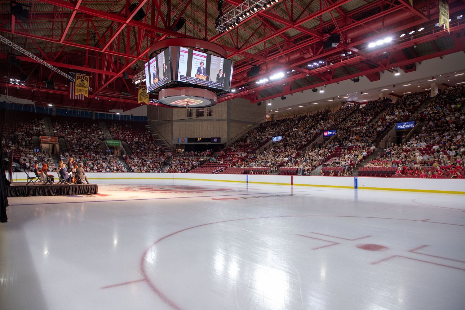A panoramic view of a brightly lit indoor ice hockey arena with red lighting and packed stadium seating.