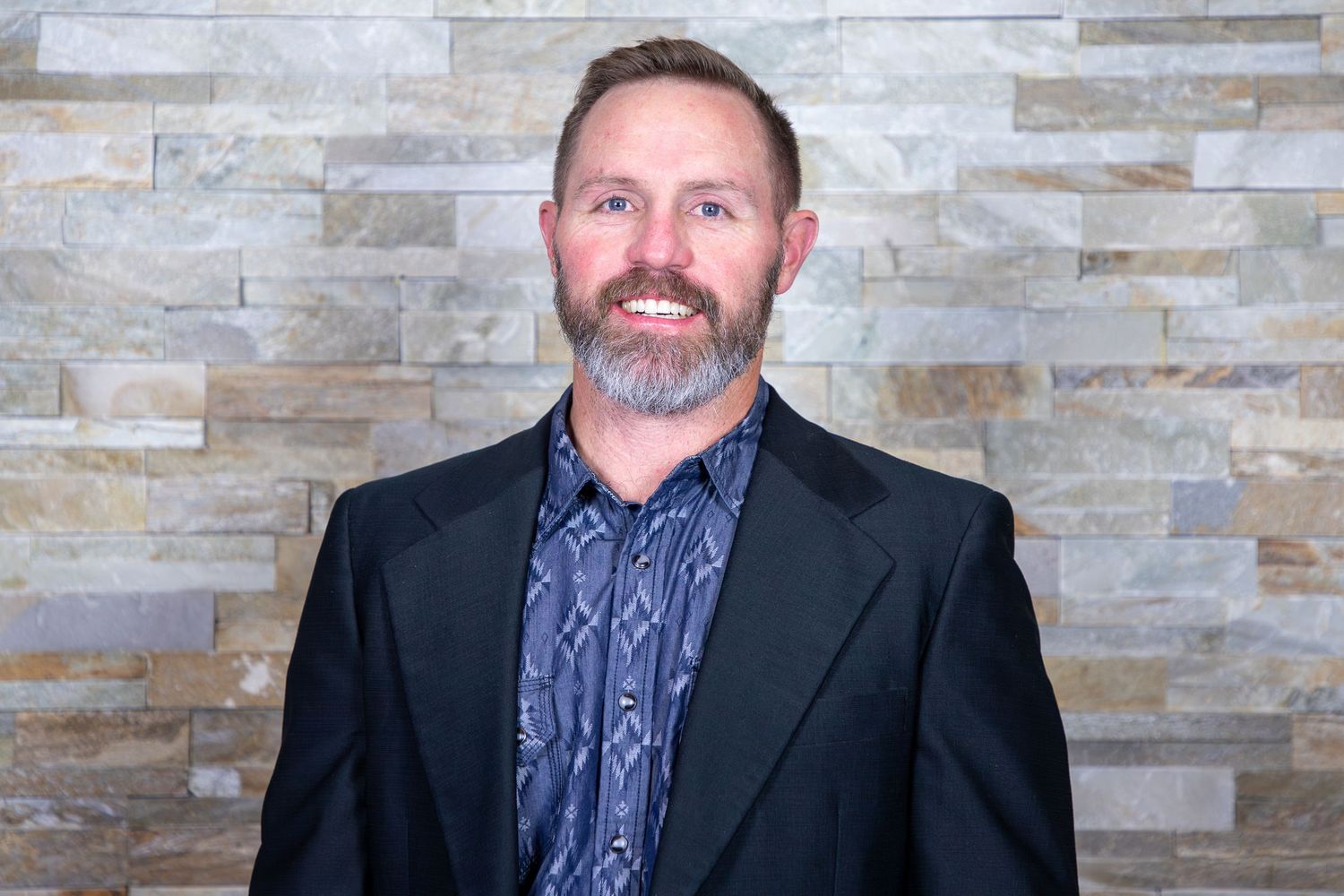 Professional headshot against stone wall backdrop wearing dark suit jacket and patterned shirt.