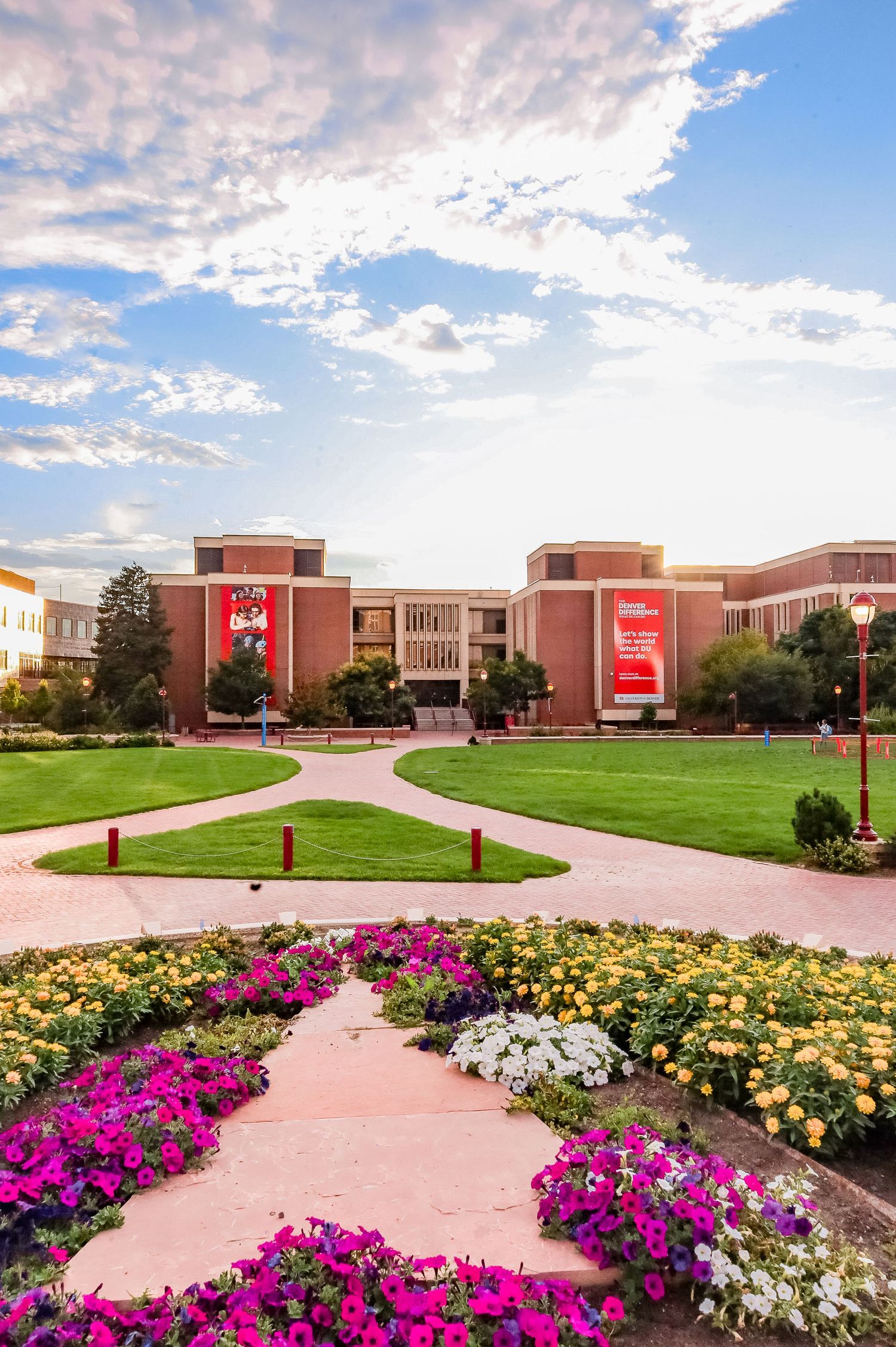 Winding pink concrete pathway through vibrant flower gardens leads to modern brick university buildings under cloudy sky.