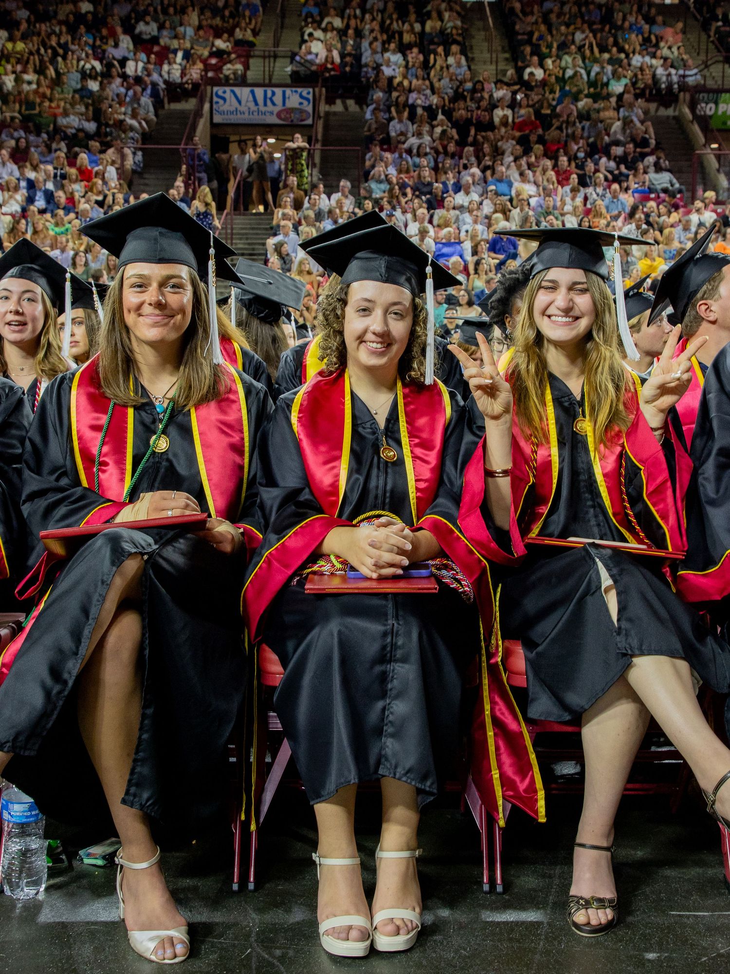 Graduates in black caps and gowns with red stoles sit together during commencement ceremony at a university.