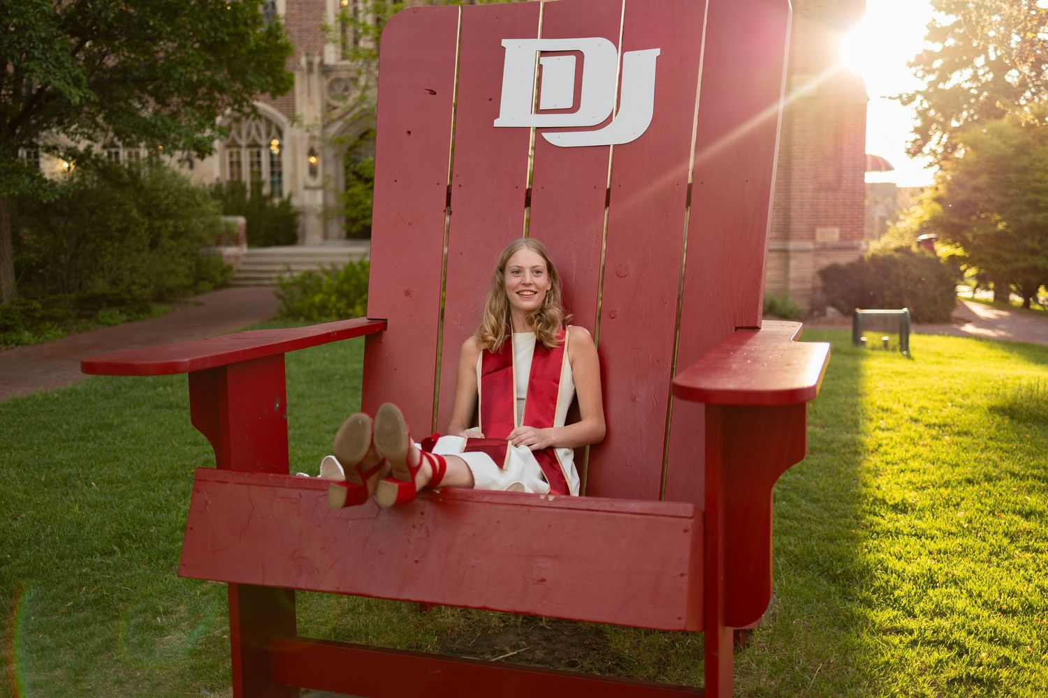 Graduate in cap and gown sitting in oversized red Adirondack chair with DU logo at University of Denver.