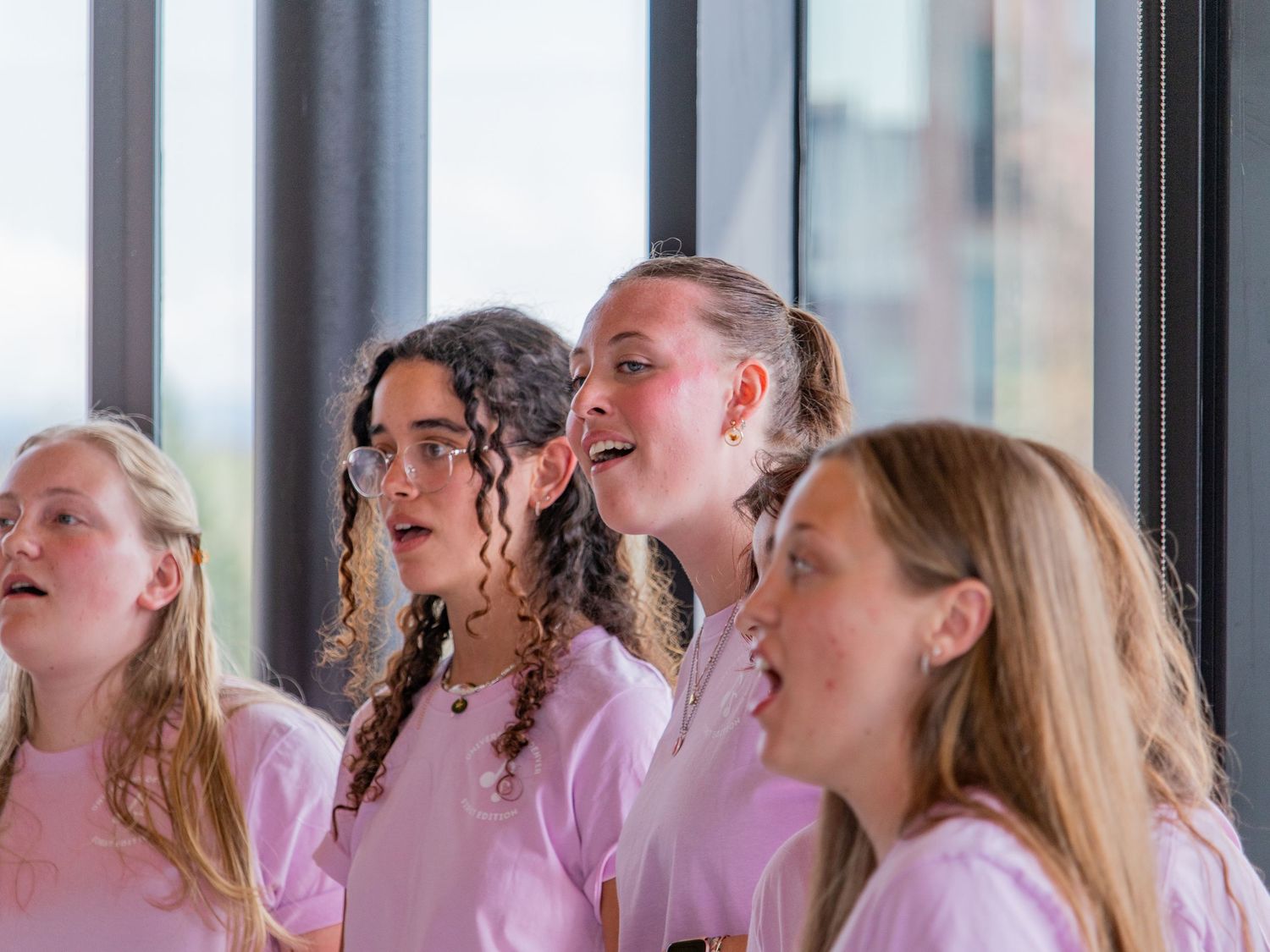 Four girls in pink shirts singing in a choir near large windows.