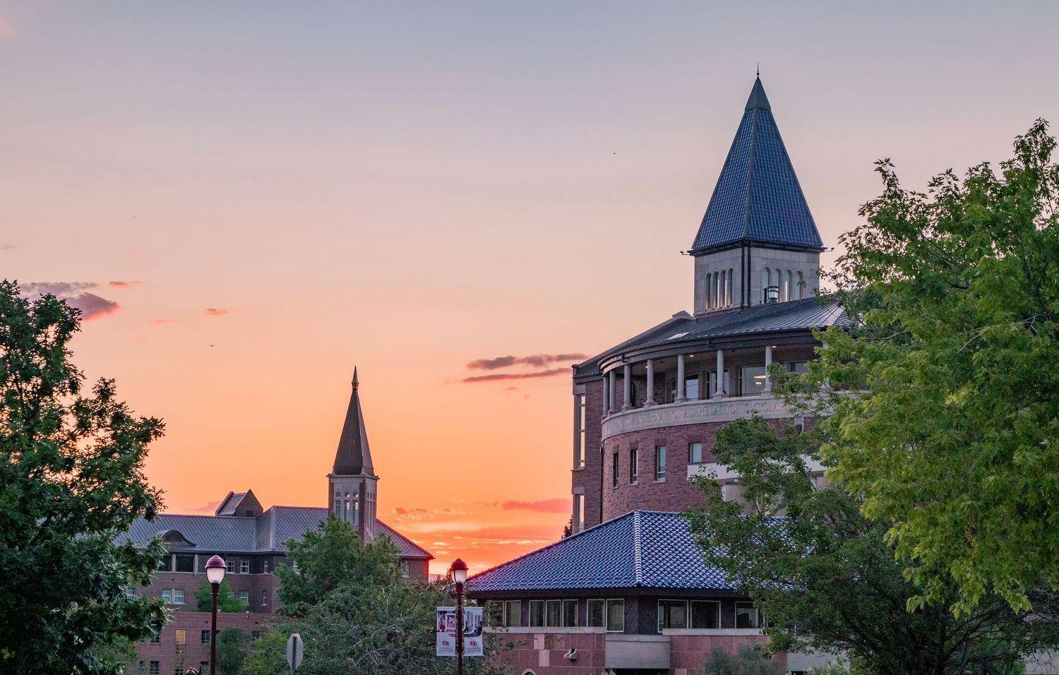 Historic brick university buildings with church steeples silhouetted against a vibrant orange and pink sunset sky.