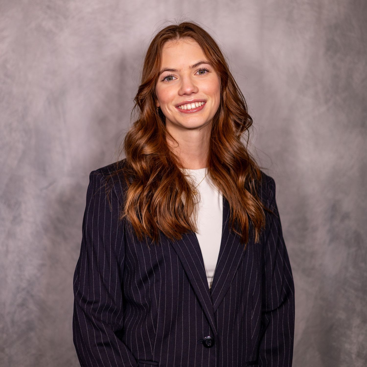 Professional headshot of a smiling young woman with auburn hair wearing a navy pinstripe blazer against a gray background.
