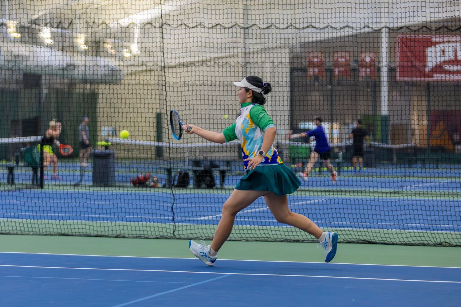 Female tennis player in green outfit mid-swing on blue indoor court during competitive match.