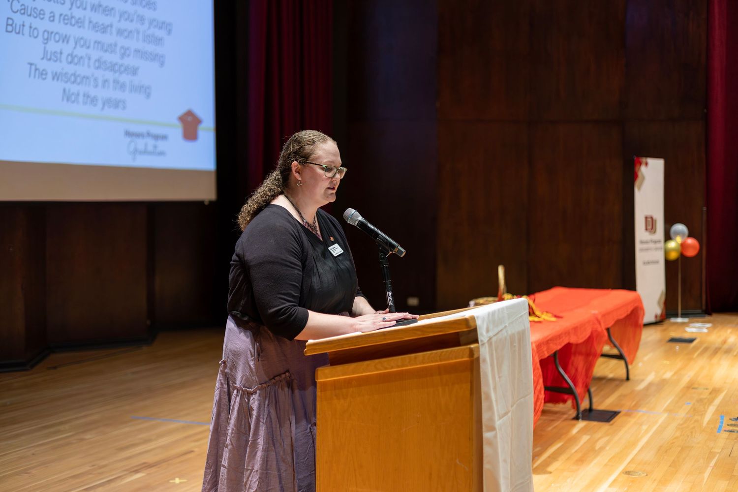 Woman speaking at podium with microphone on auditorium stage with projection screen and red table in background.