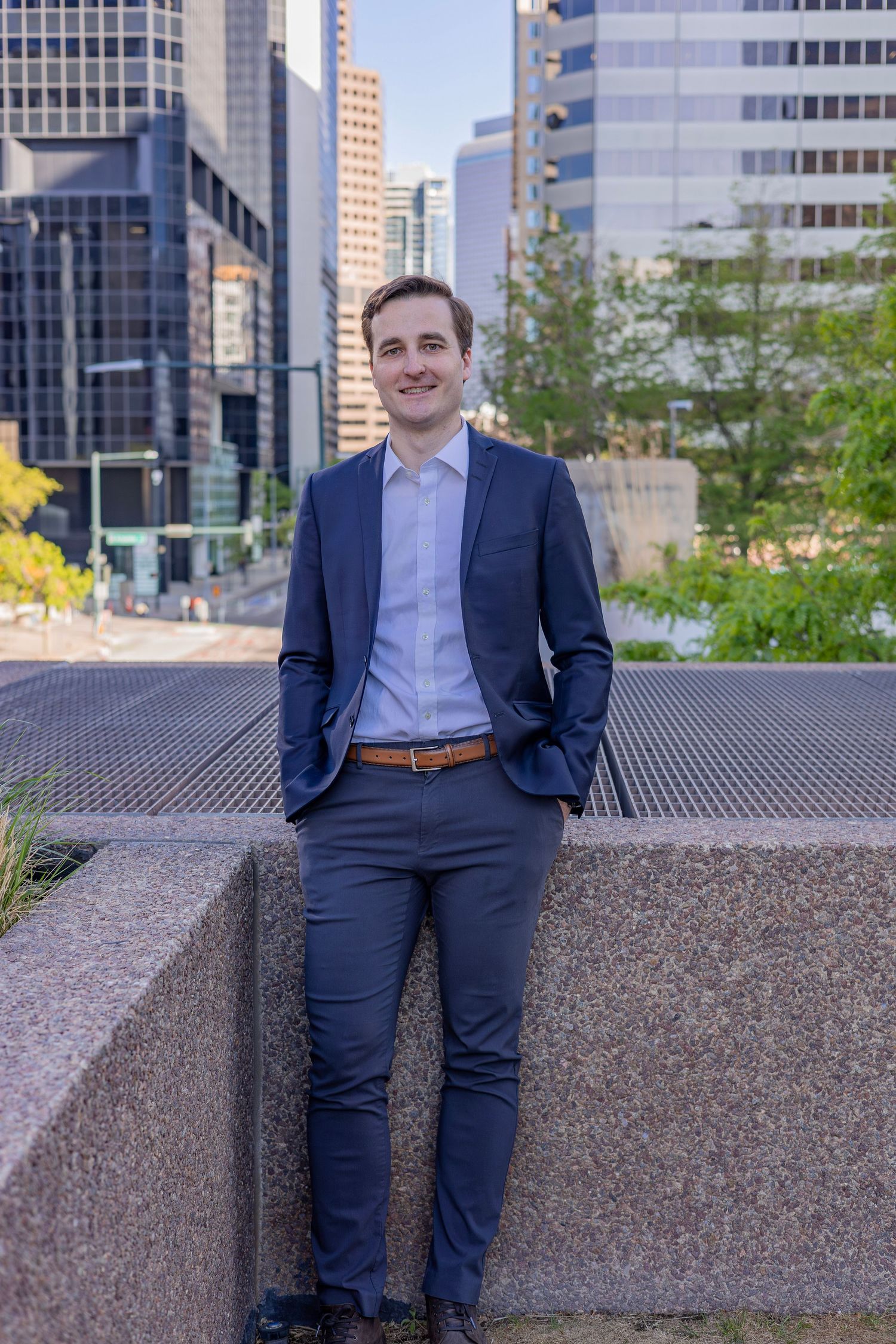 Young professional man in navy suit smiling outdoors with city skyscrapers in background.