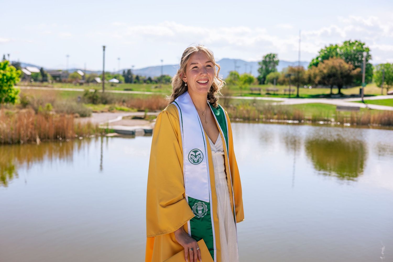 Smiling graduate in yellow and green cap and gown poses outdoors near a pond on a sunny day.