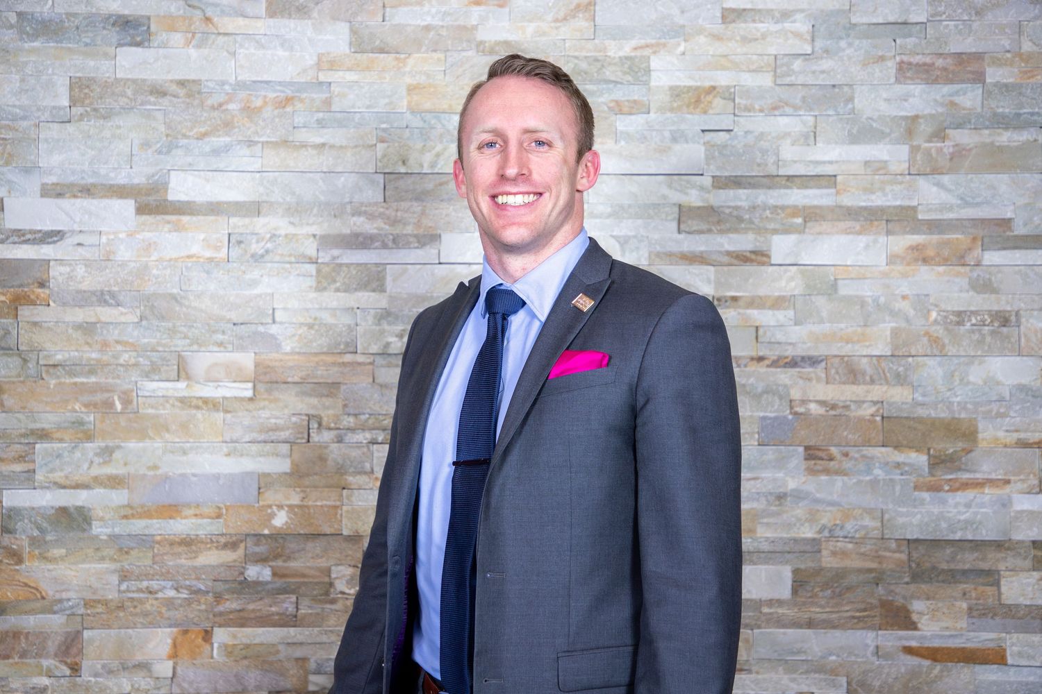 Professional wearing a dark gray suit and pink boutonniere stands against a textured stone wall backdrop.