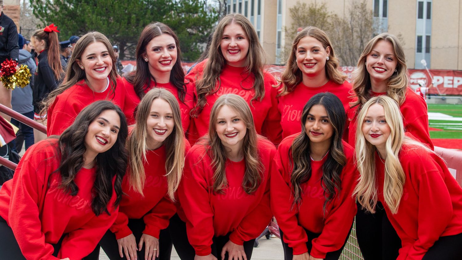 Group of ten young women in matching red sweatshirts posing together outdoors at a sports field event.