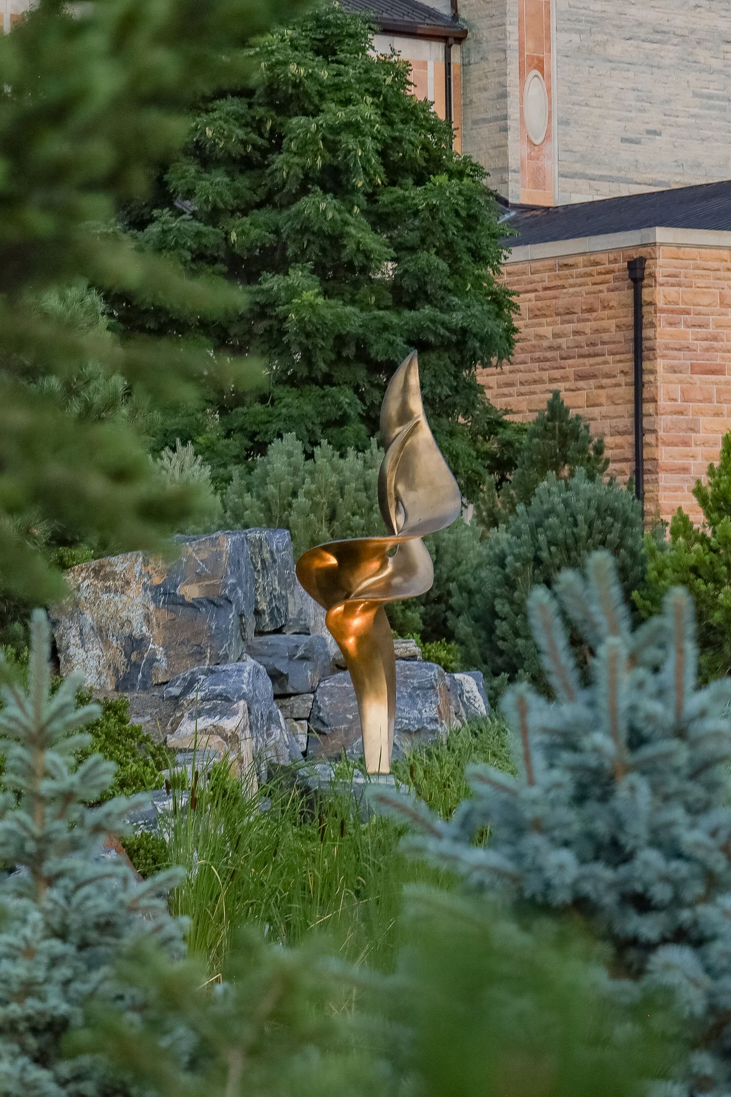 Golden abstract sculpture amid lush evergreen garden with stone wall and brick building backdrop.