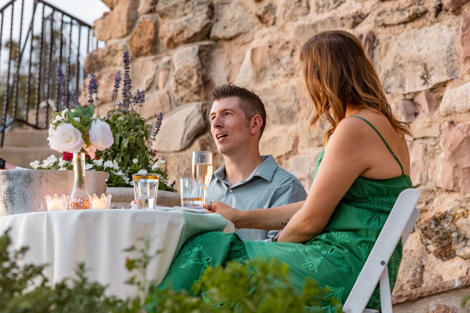 Couple enjoying a romantic outdoor candlelit dinner by a stone wall, smiling and sharing champagne.