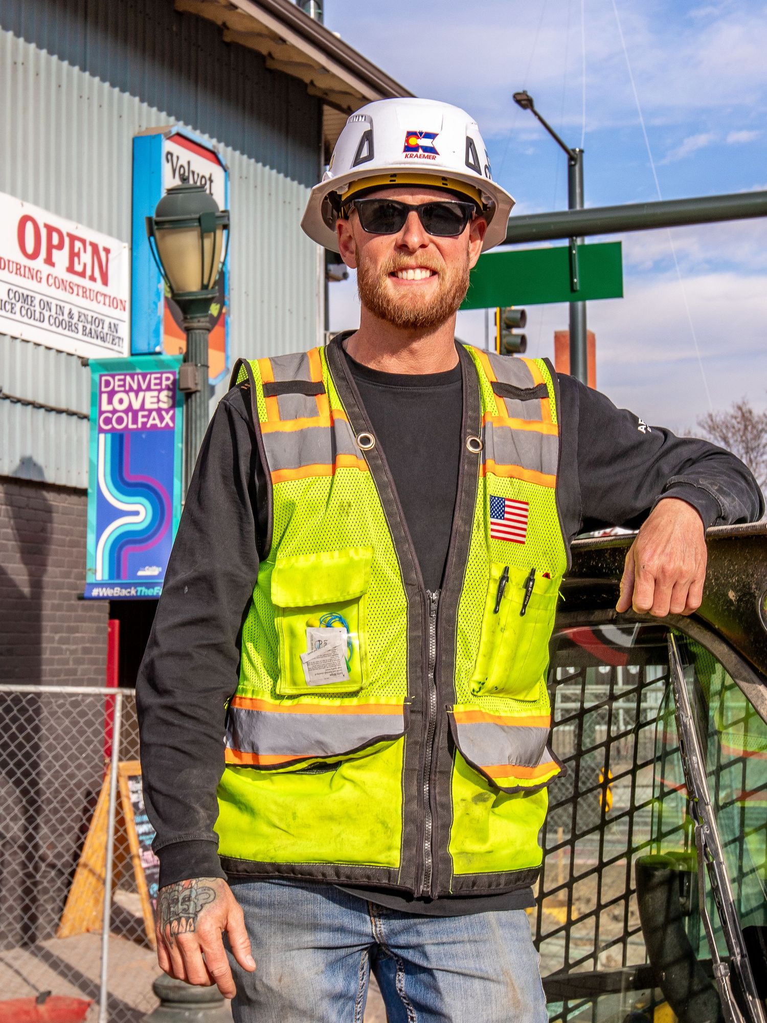 Construction worker in hi-vis vest and hard hat smiles near Denver Loves Colfax sign on a sunny day.