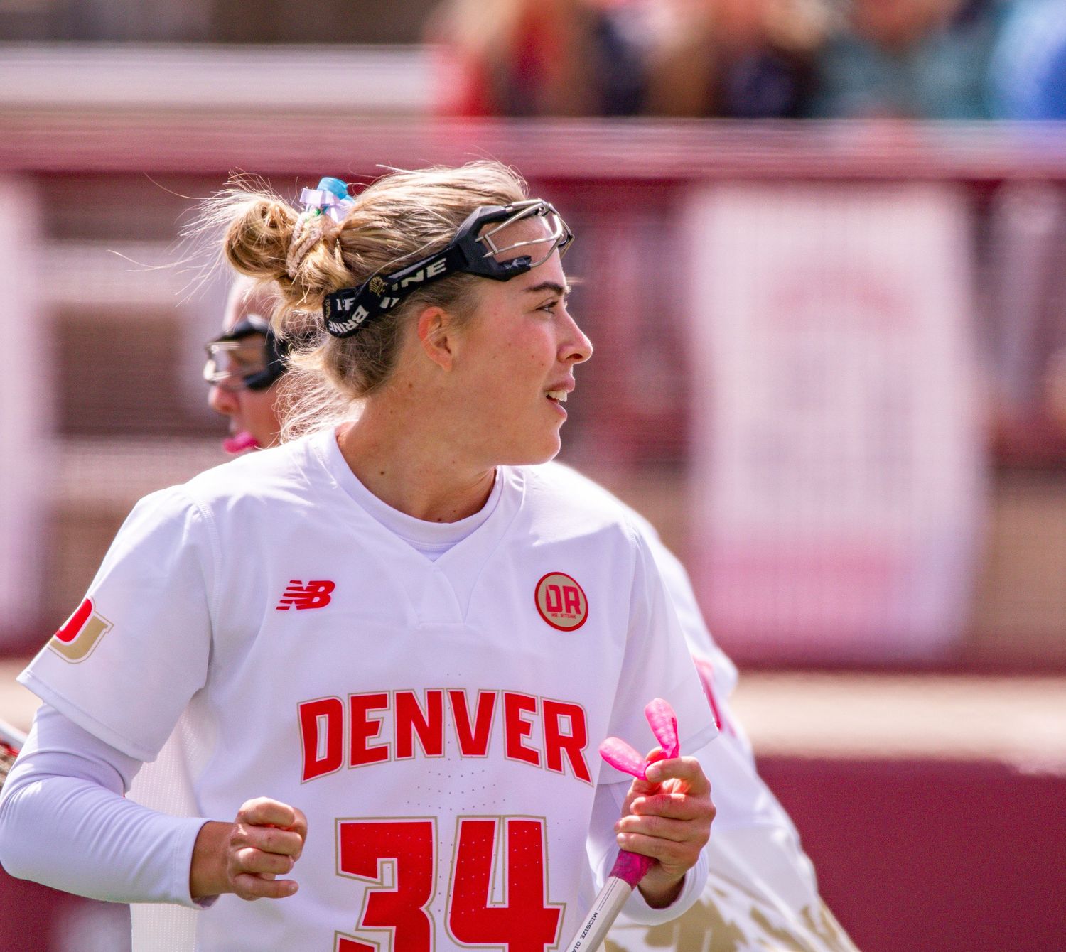 College lacrosse player in white Denver jersey number 34 runs during competitive match at stadium.